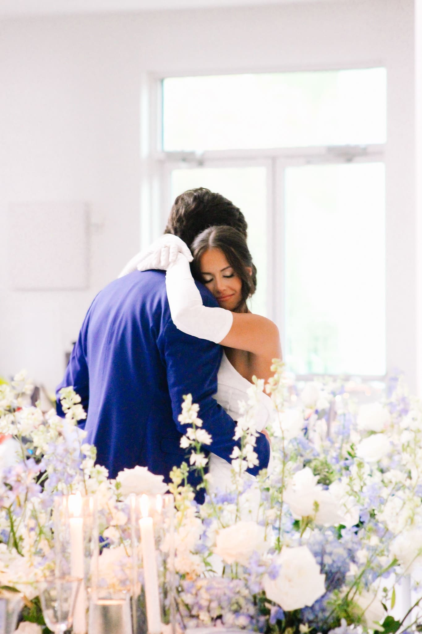 A bride and groom embrace tenderly indoors, surrounded by soft blue and white flowers and lit candles, with natural light streaming through a window in the background.