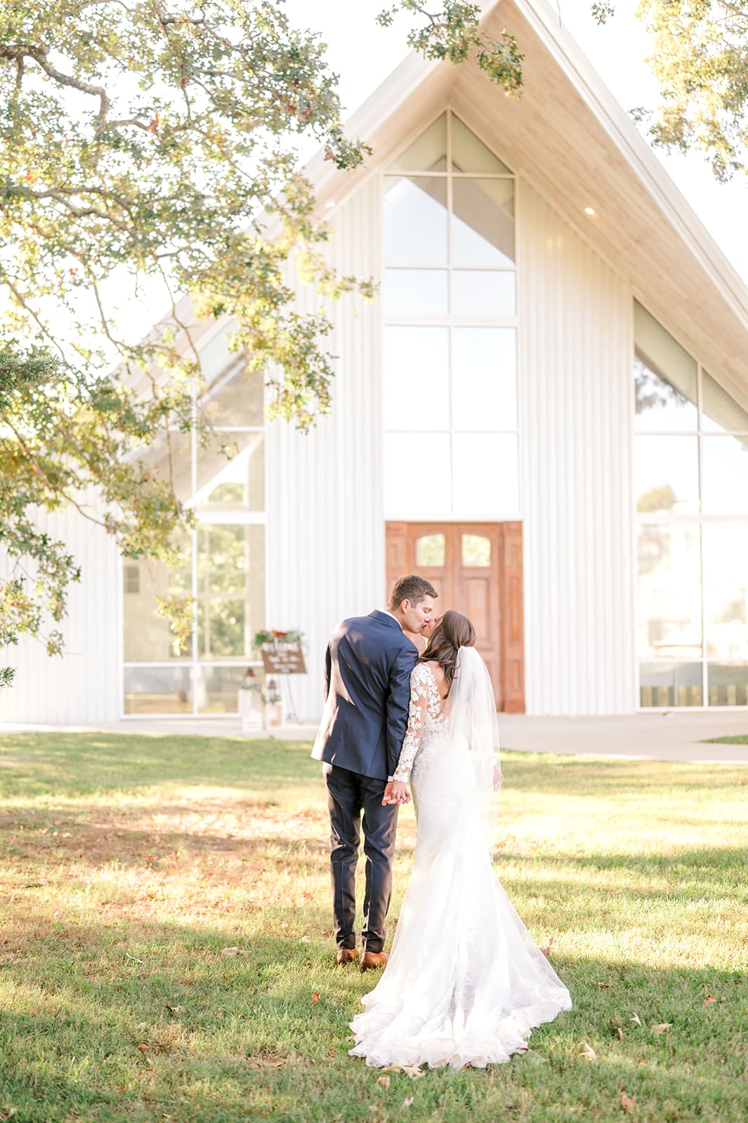 A bride and groom stand outside a modern white chapel, facing away and holding hands, with sunlight filtering through the trees on a grassy lawn. The bride wears a long white gown and veil; the groom wears a dark suit.