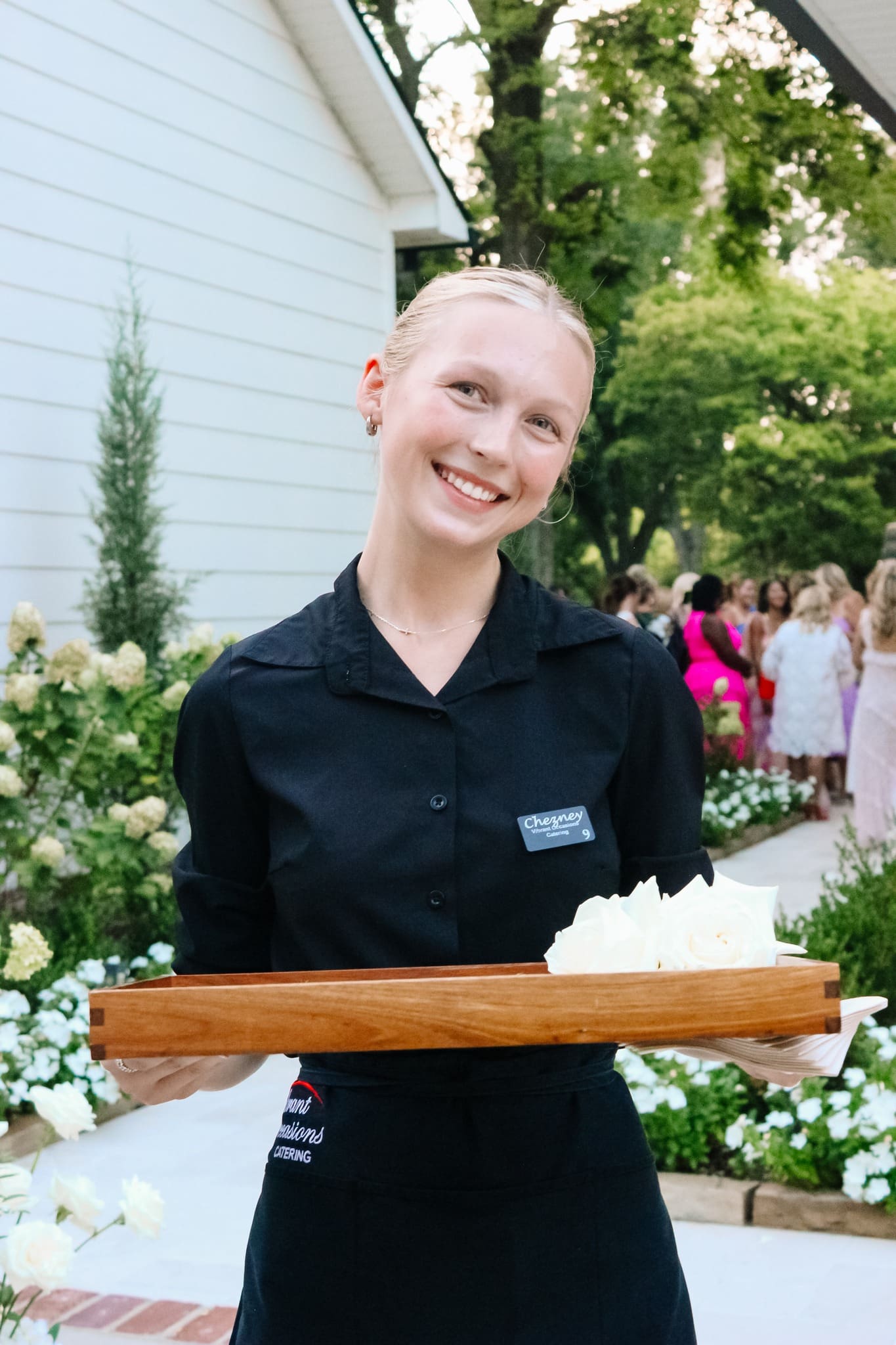 Staff member serving appetizers A smiling server in a black uniform holds a wooden tray with white napkins at an outdoor event, with guests and greenery visible in the background.