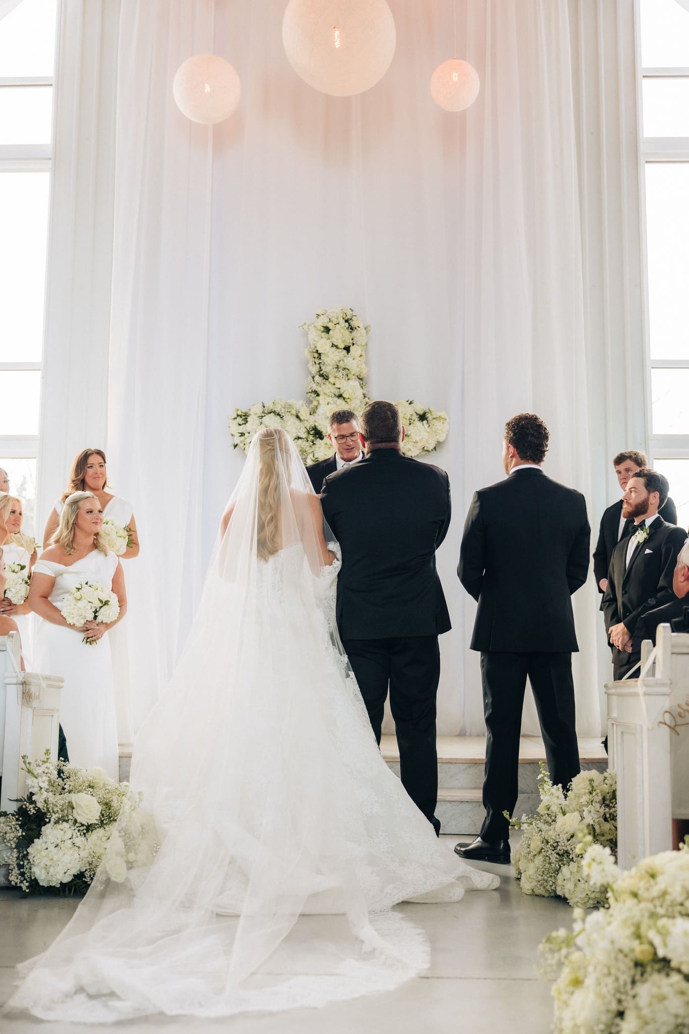 A bride in a long white gown and veil stands at the altar with two men in black suits. Bridesmaids and groomsmen stand nearby. The setting is decorated with white flowers and a large floral cross.