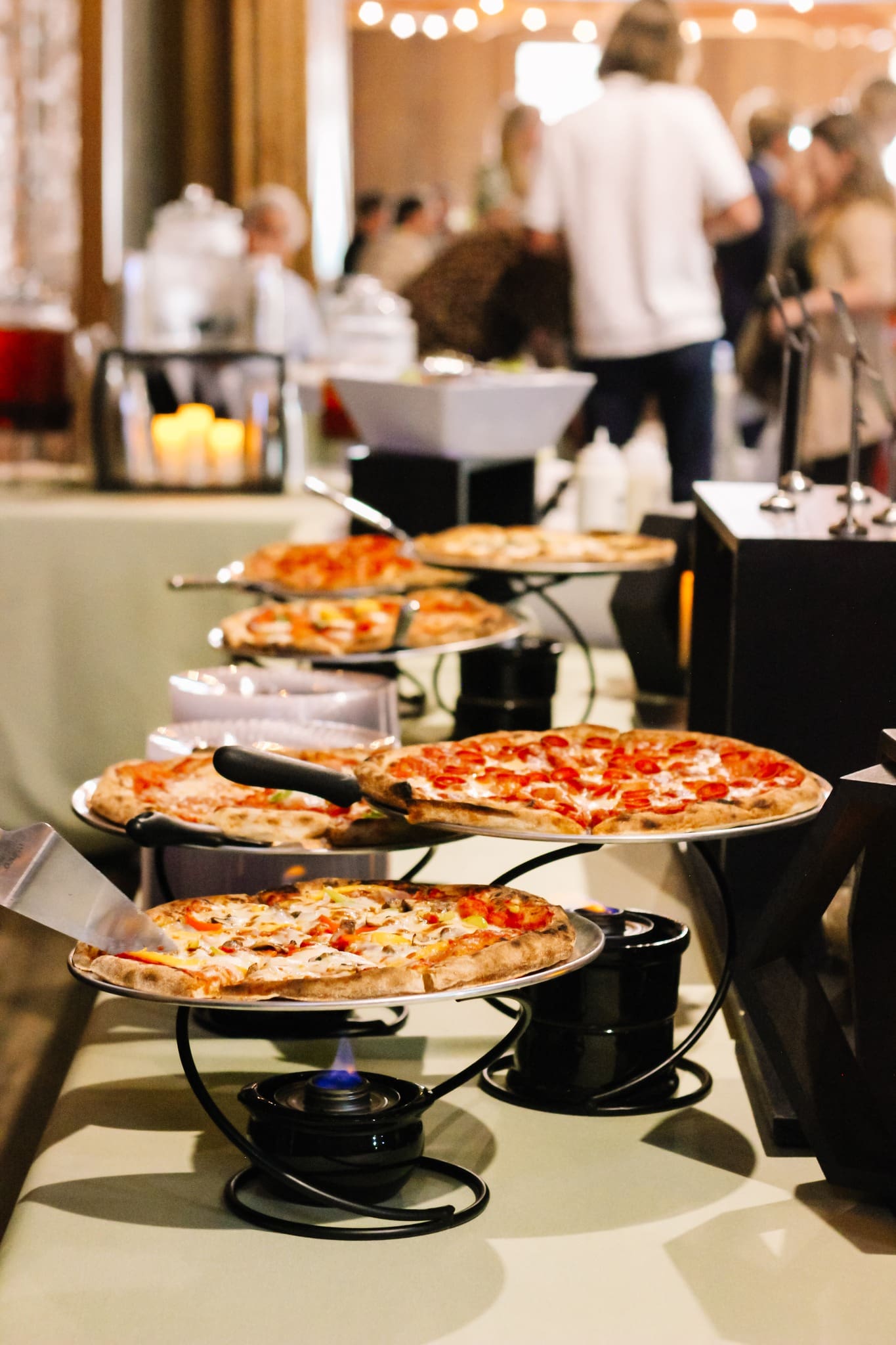 Pizza Action Station A buffet table displays several pizzas on elevated stands, with people blurred in the background at what appears to be a social gathering or event.