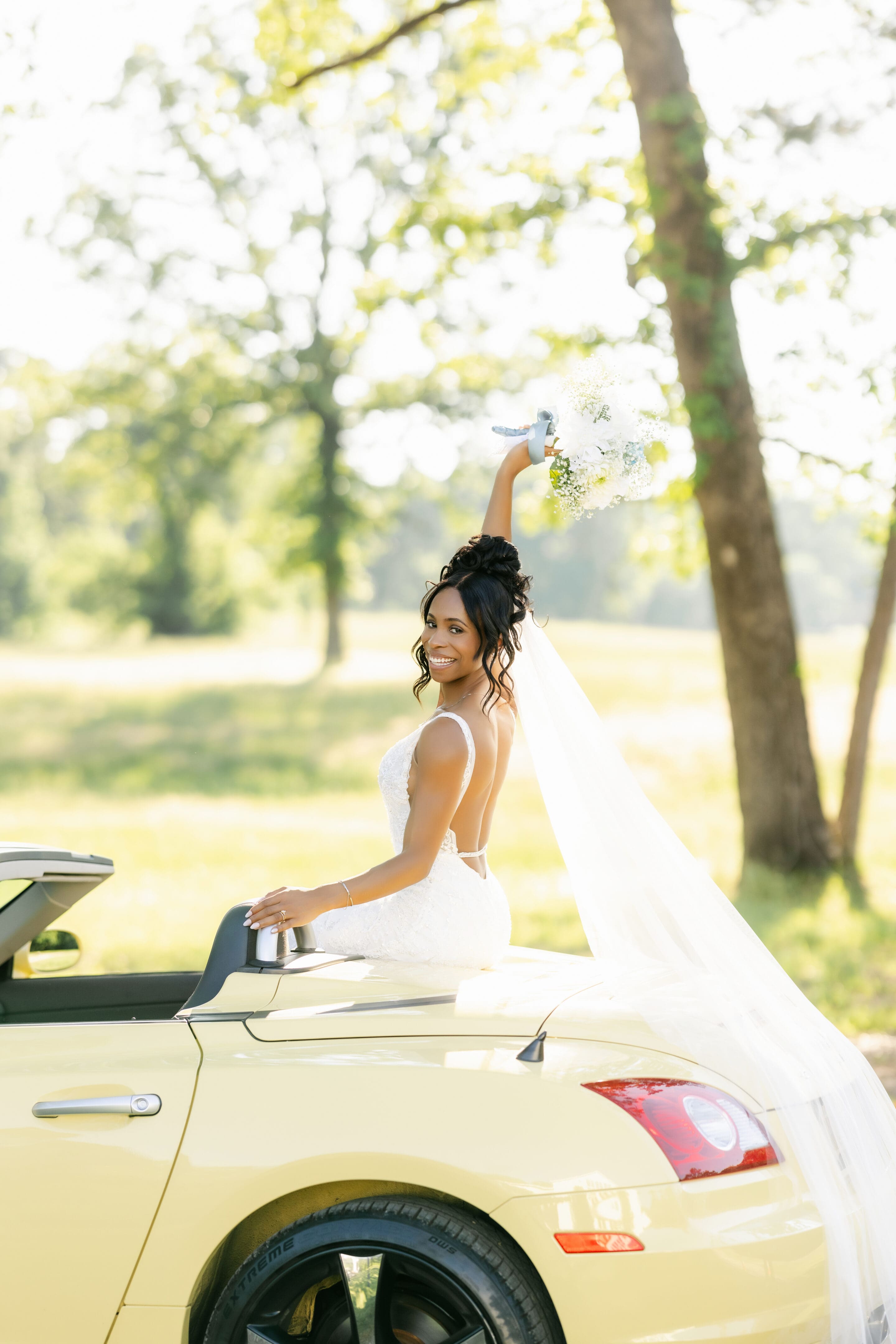 A bride in a lace wedding dress holds a vibrant bouquet with red, pink, and dark flowers, and green foliage, outdoors with greenery in the background. Her face is partially visible.