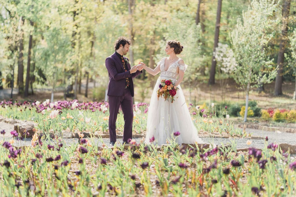A bride in a white dress holding a bouquet and a groom in a purple suit hold hands and smile at each other during their enchanting Eclipse Weekend Wedding, surrounded by blooming flowers and lush greenery in the garden.