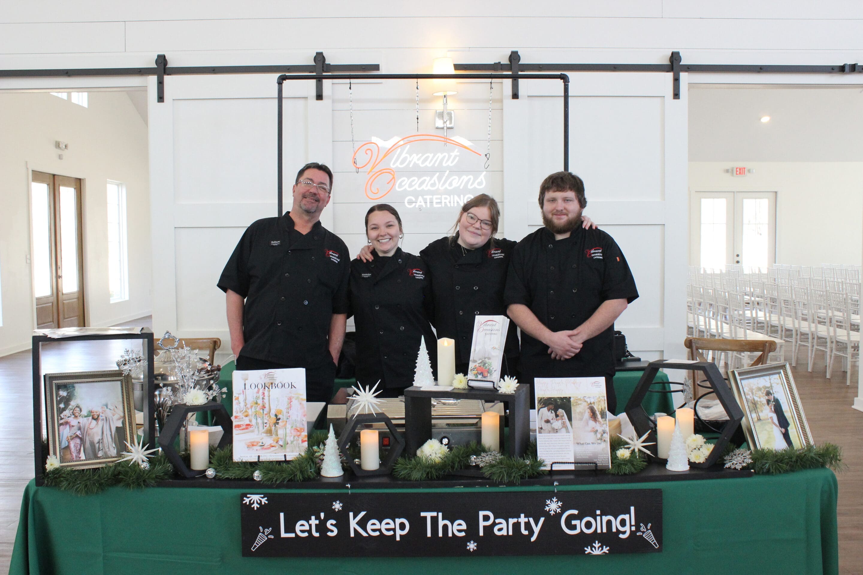Four catering staff in black uniforms stand behind a decorated table with candles, photos, and a sign that reads Let’s Keep The Party Going! The backdrop displays a Vibrant Occasions Catering sign at this Married & Bright Wedding Event.
