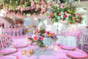 A vibrant wedding reception table features a floral centerpiece with pink, blue, and purple flowers, surrounded by candles. In the background, large floral arrangements and hanging flowers decorate the bright, elegant venue.
