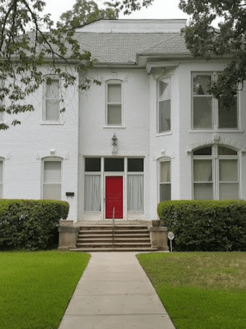 The Gaines House 16th Awards Dinner Menu A white, two-story house with large windows and a bright red front door, ideal for social and corporate catering events, sits behind a green lawn and concrete walkway, accessed by steps flanked by trimmed bushes.