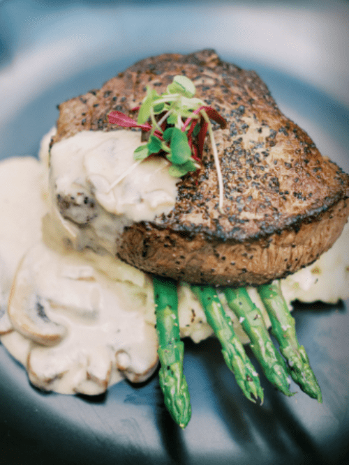 2025 Wedding Catering Trends Perfect for social and corporate catering, this grilled steak is topped with microgreens and served on mashed potatoes with creamy mushroom sauce and asparagus spears, elegantly presented on a dark plate.
