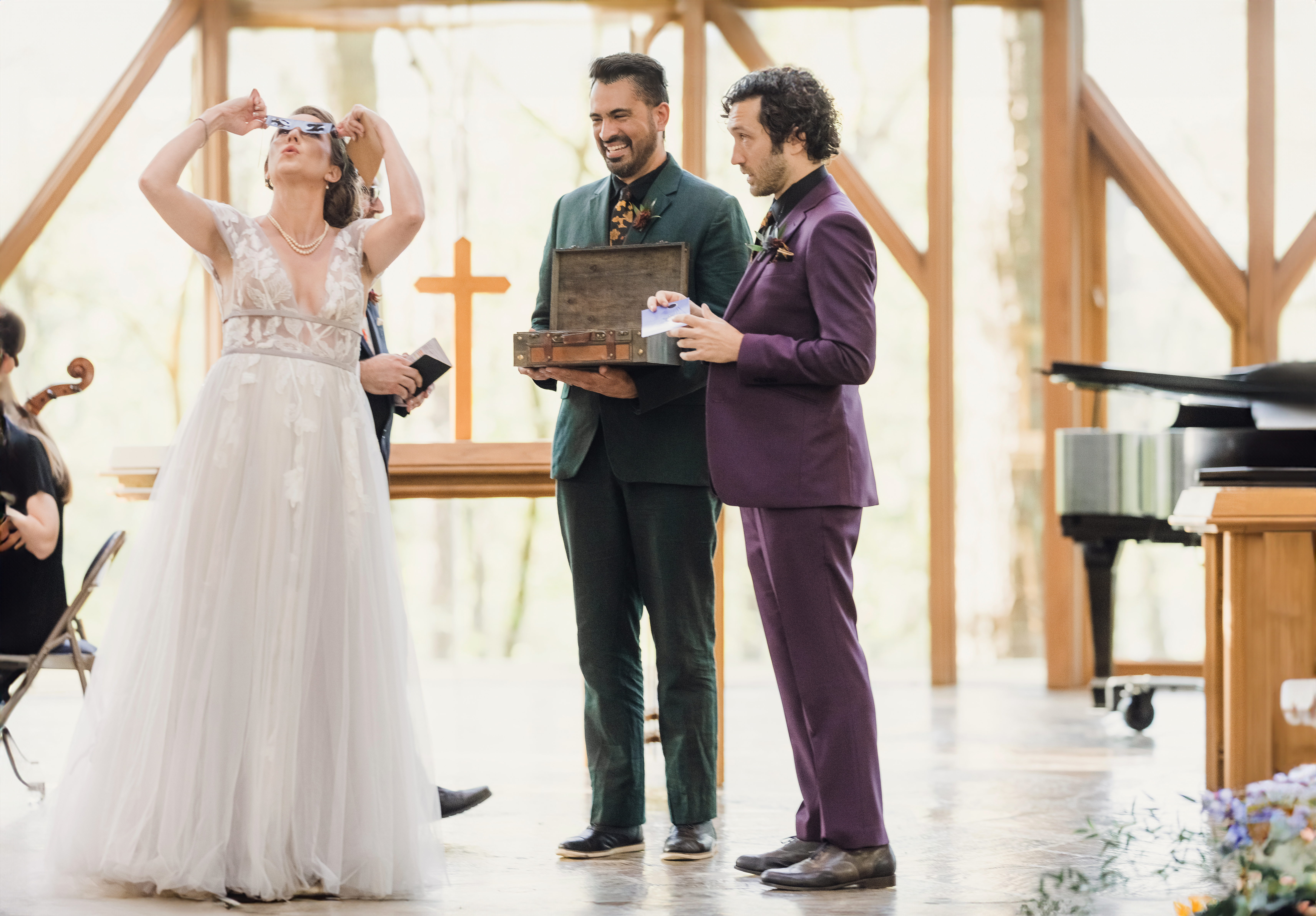 A bride in a white gown is laughing and holding up sunglasses during what appears to be a wedding ceremony, while two grooms in dark suits stand beside her, one holding a wooden box and smiling. The setting seems to be indoors with large windows and wooden beams, suggesting a rustic or chapel-like venue.