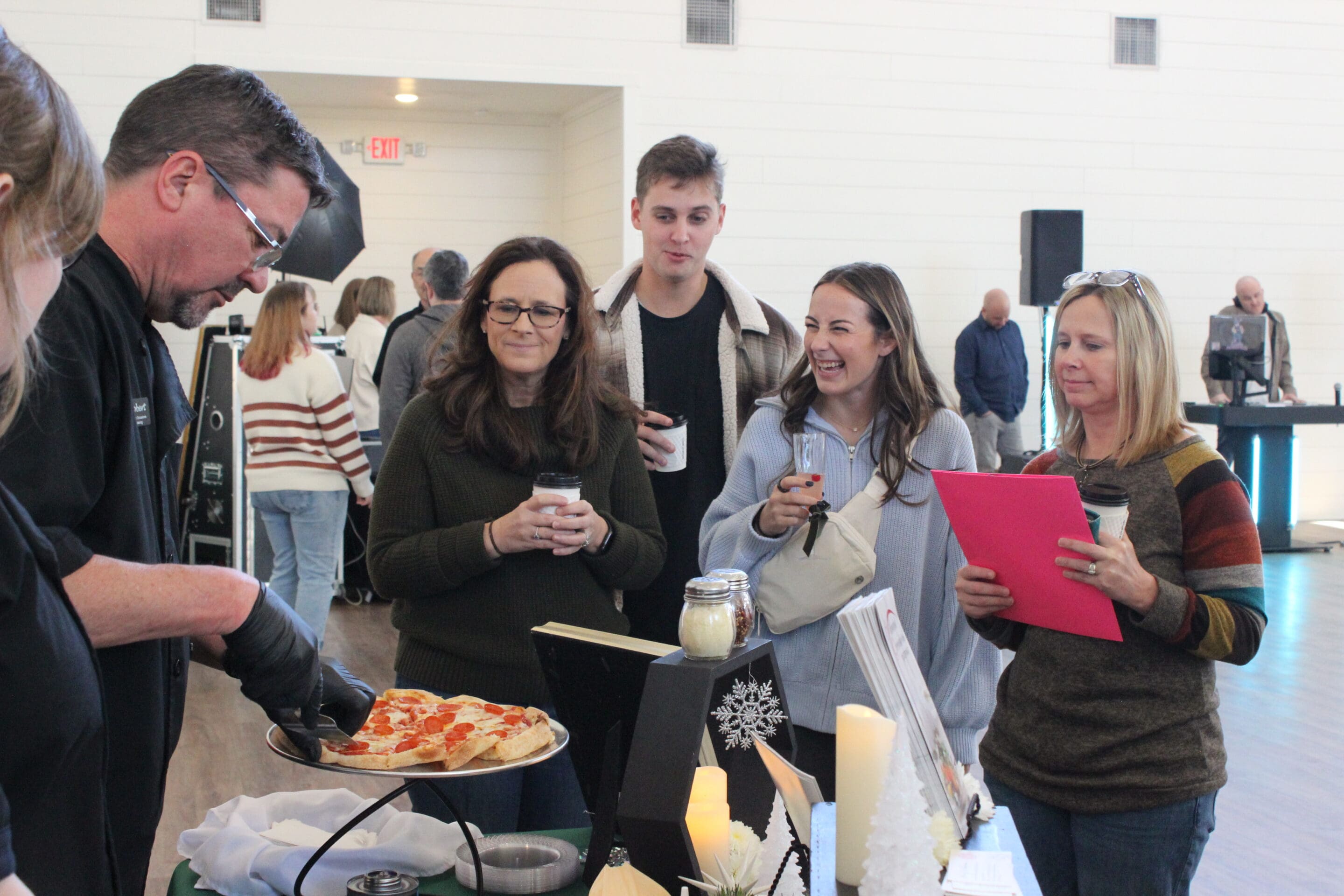 At a Married & Bright Wedding Event, a man serves pizza to four smiling women and one man. The group holds coffee cups and papers, with other guests and festive decorations visible in the background.