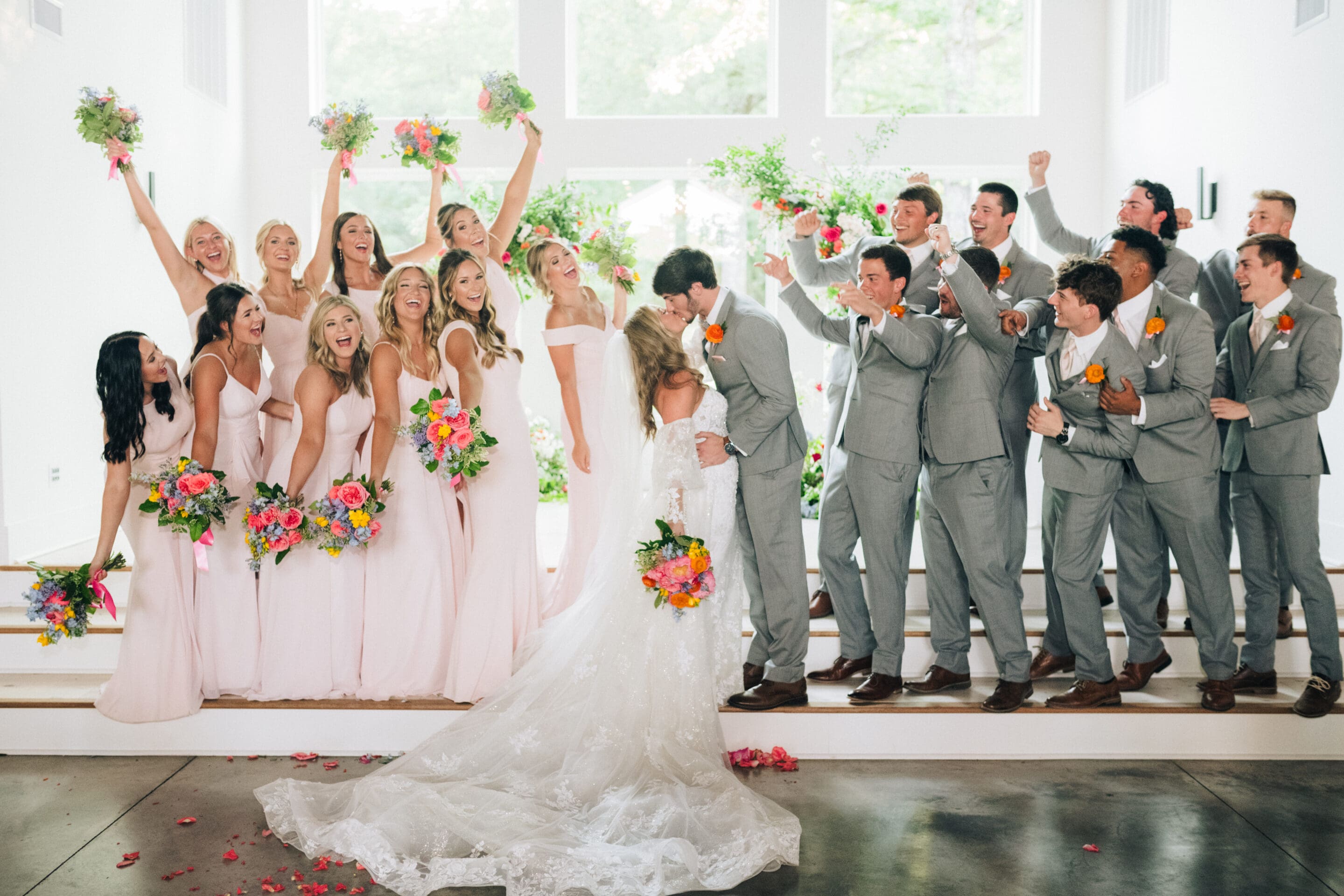 A bride and groom kiss surrounded by bridesmaids in pale pink dresses and groomsmen in gray suits, all cheering and holding colorful flower bouquets in a bright, modern venue.