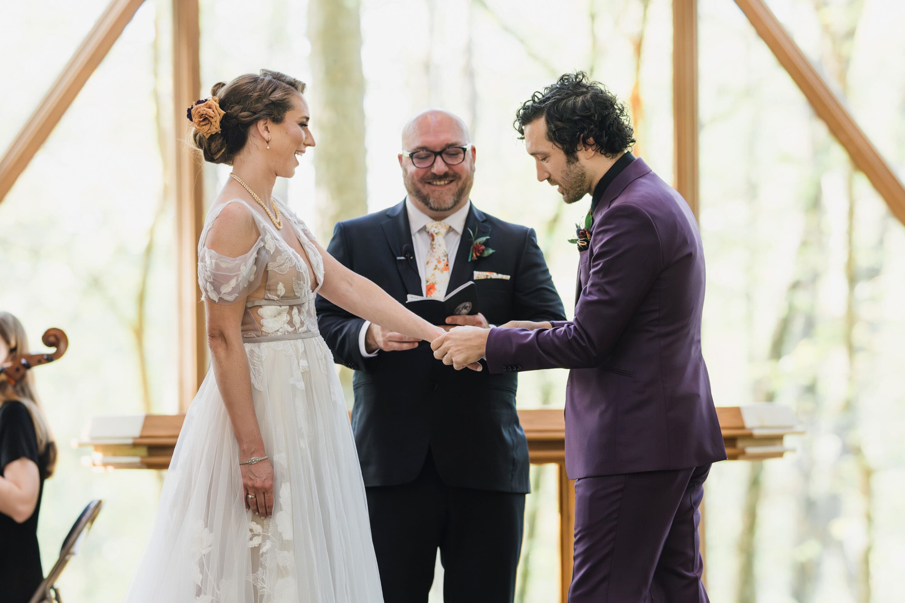 A bride and groom stand facing each other holding hands during their Eclipse Weekend Wedding ceremony, with an officiant smiling behind them inside a bright, sunlit venue with large windows.