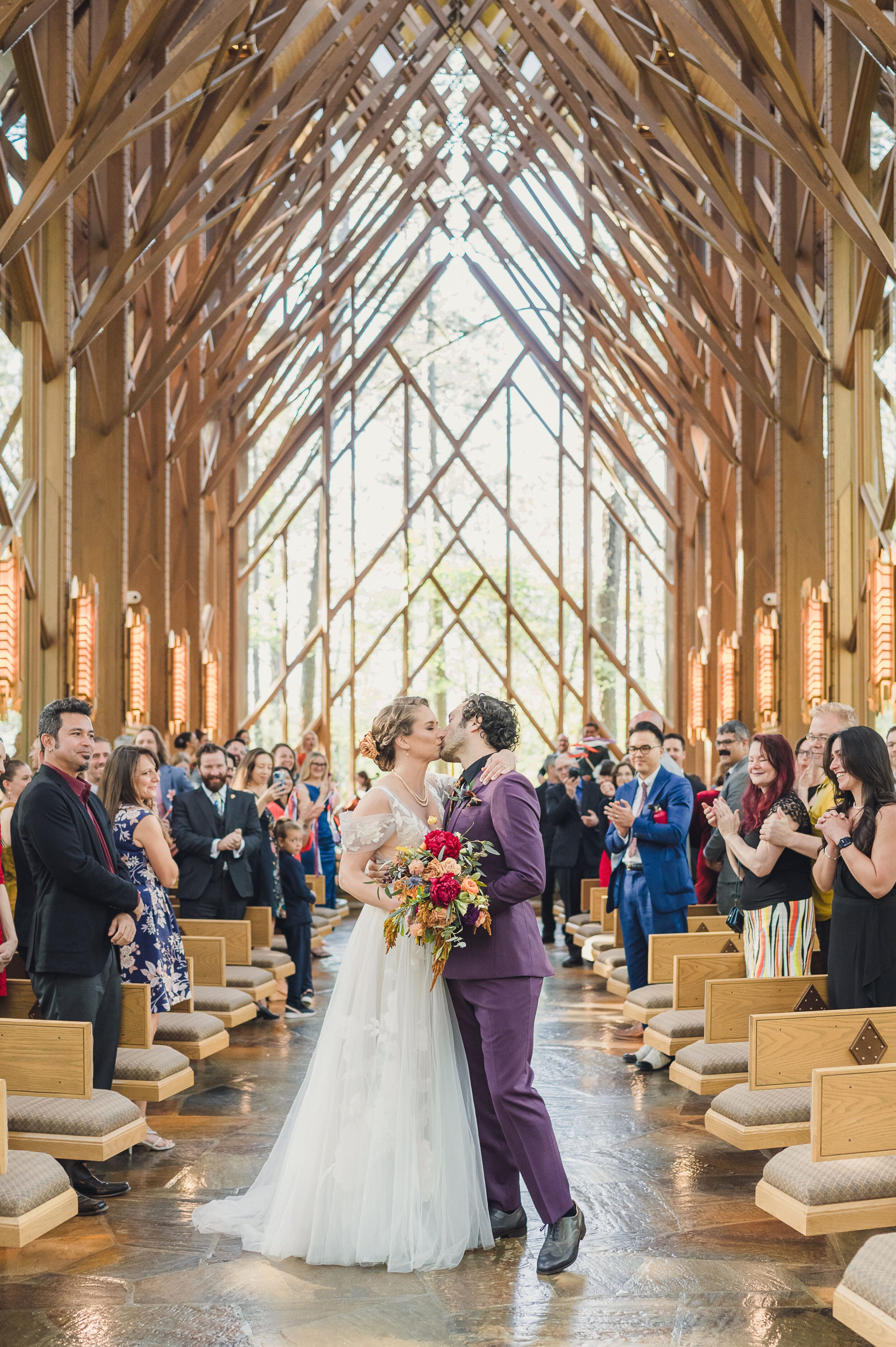 A newlywed couple kisses while walking down the aisle of a beautifully lit wooden chapel during their Eclipse Weekend Wedding. Guests stand on either side, smiling and applauding, as sunlight streams through large windows above.