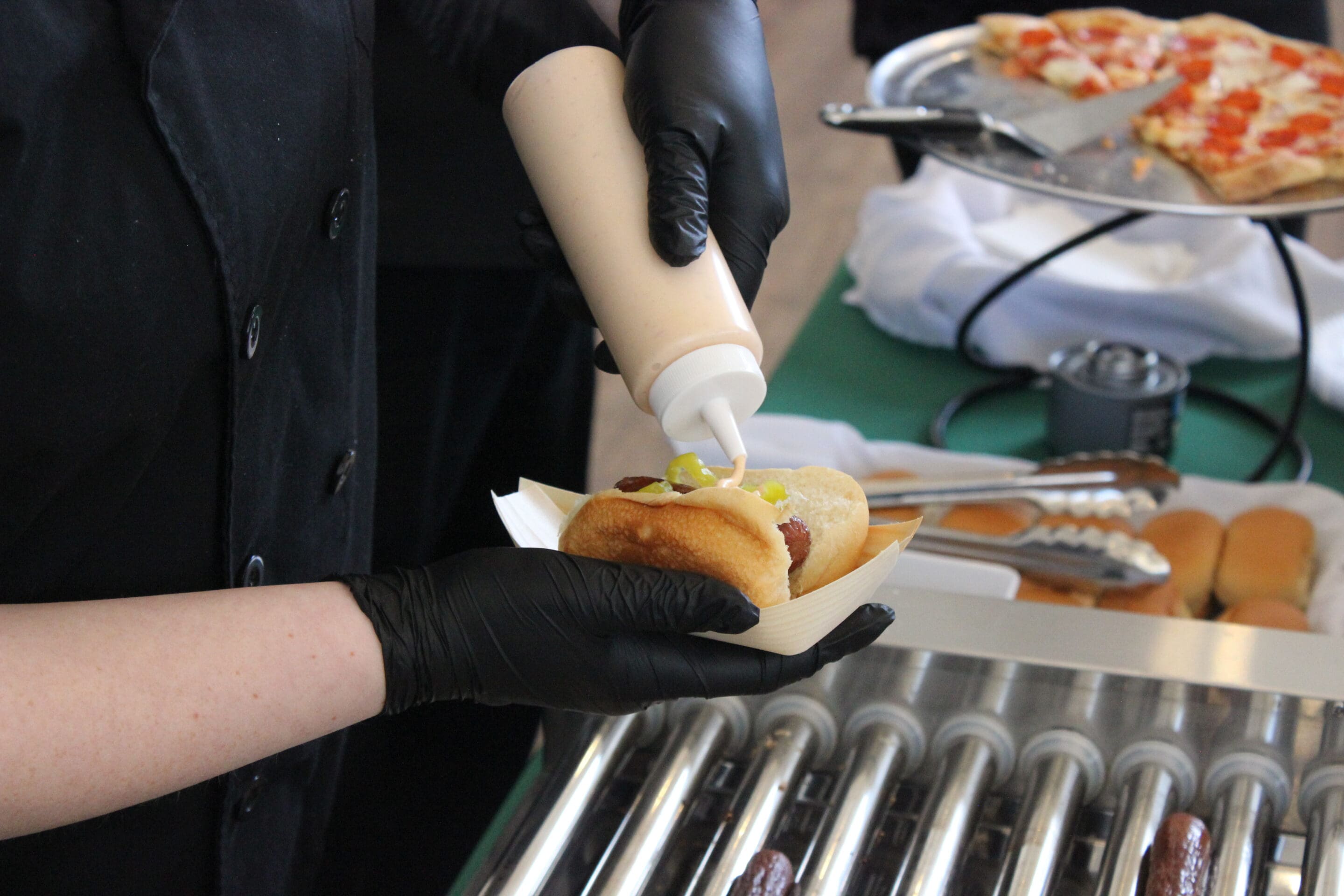 At a Married & Bright Wedding Event, a person wearing black gloves squeezes sauce onto a hot dog in a bun, with more hot dogs on a roller grill and a plate of pizza slices visible in the background.