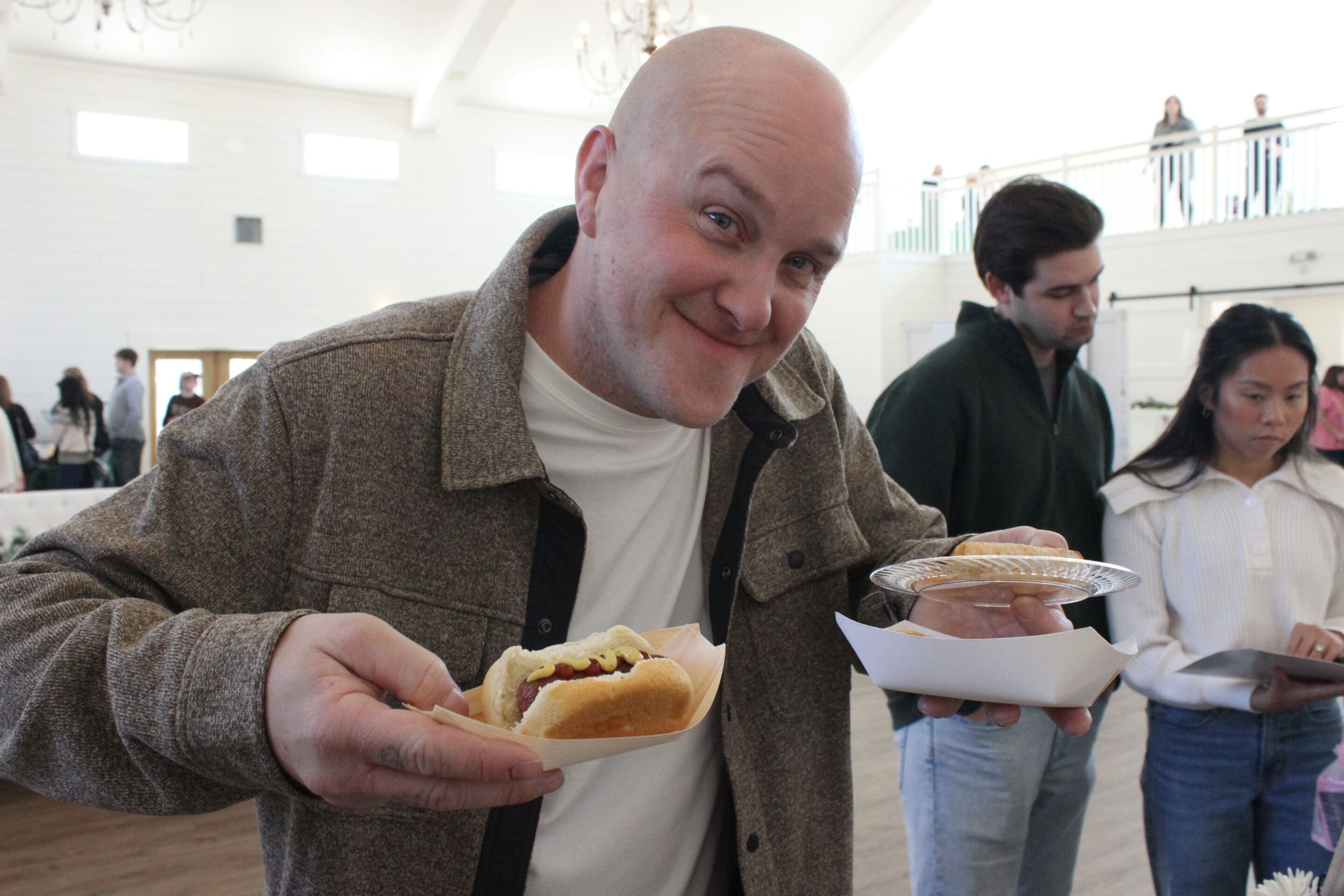 At a Married & Bright Wedding Event, a smiling bald man in a brown jacket holds a hot dog and a pie slice, enjoying the indoor celebration while others around him serve themselves food.