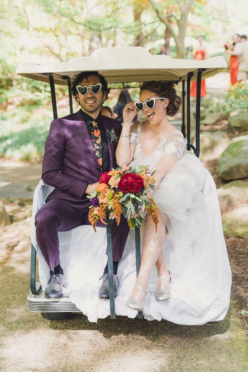 A bride and groom wearing heart-shaped sunglasses sit on a golf cart during their Eclipse Weekend Wedding. The groom’s in a purple suit with a floral tie, and the bride lifts her shades, both grinning joyfully outdoors with her vibrant bouquet.