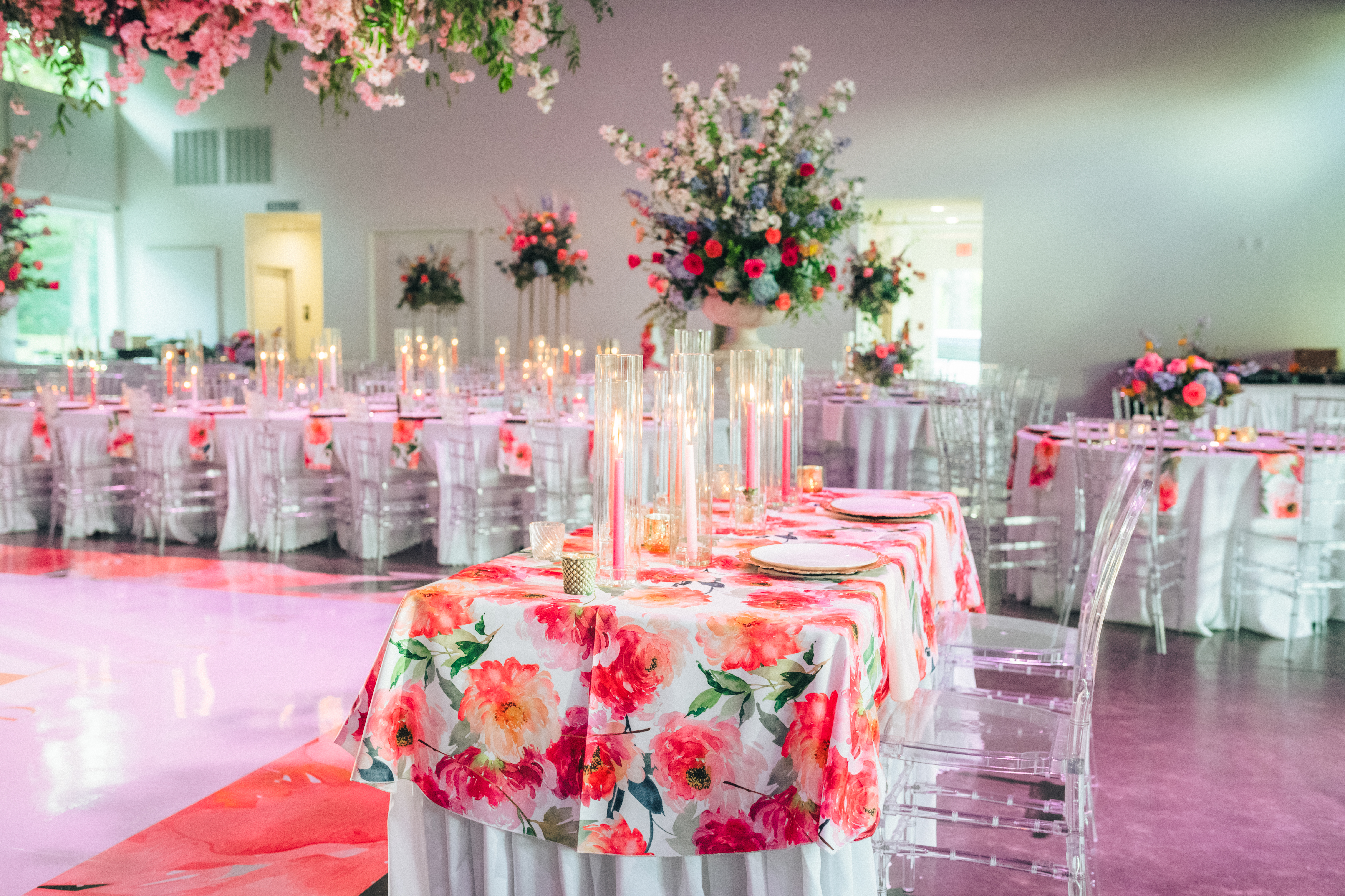 The image shows an elegantly decorated indoor event space, possibly for a wedding reception, with round tables covered in floral tablecloths and adorned with tall floral centerpieces and candles in glass cylinders. Clear acrylic chairs surround the tables, and floral decorations hang from the ceiling.