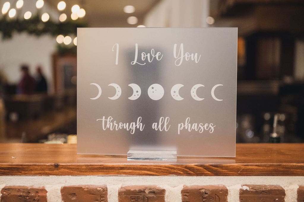 A frosted acrylic sign on a wooden ledge displays “I Love You through all phases” with moon phase illustrations—perfect for an Eclipse Weekend Wedding. The softly blurred background adds a romantic touch.