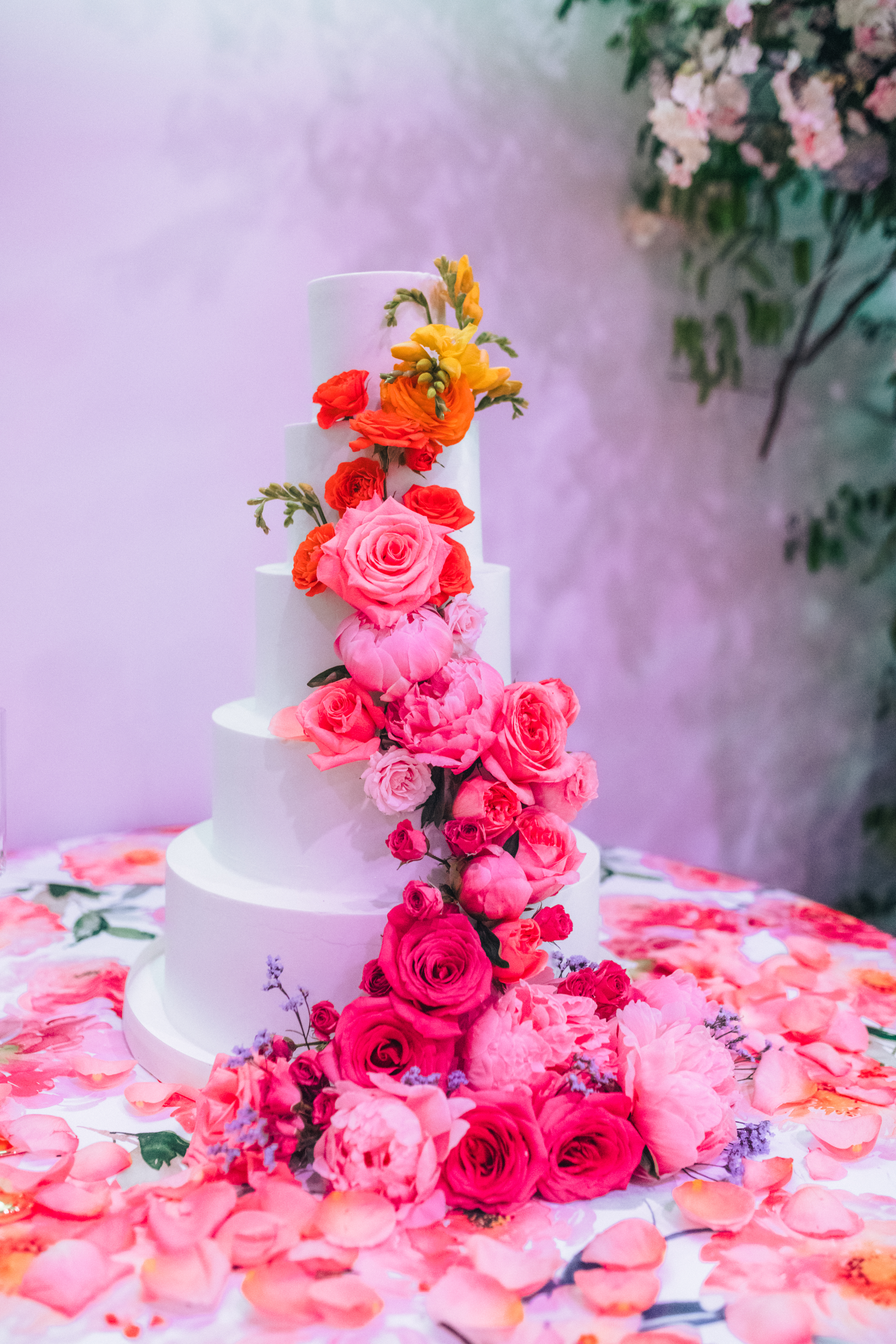 The image shows a three-tiered white wedding cake lavishly decorated with a cascade of vibrant pink, red, and orange roses and other flowers. The cake sits on a table covered with a floral patterned tablecloth and scattered petals.