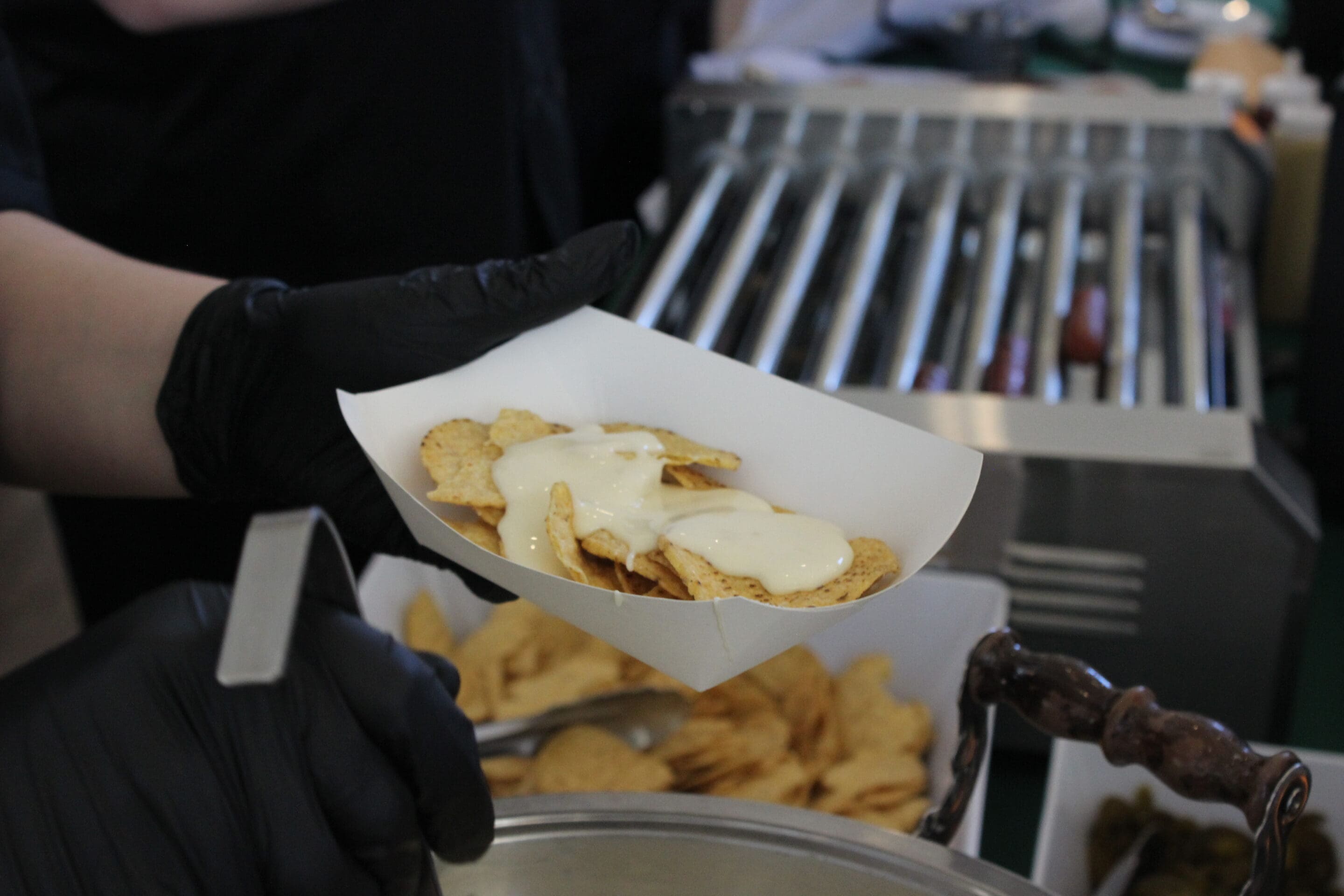 At a Married & Bright Wedding Event, a person wearing black gloves holds a paper tray filled with tortilla chips smothered in white cheese sauce, with more chips and a hot dog roller seen in the background.