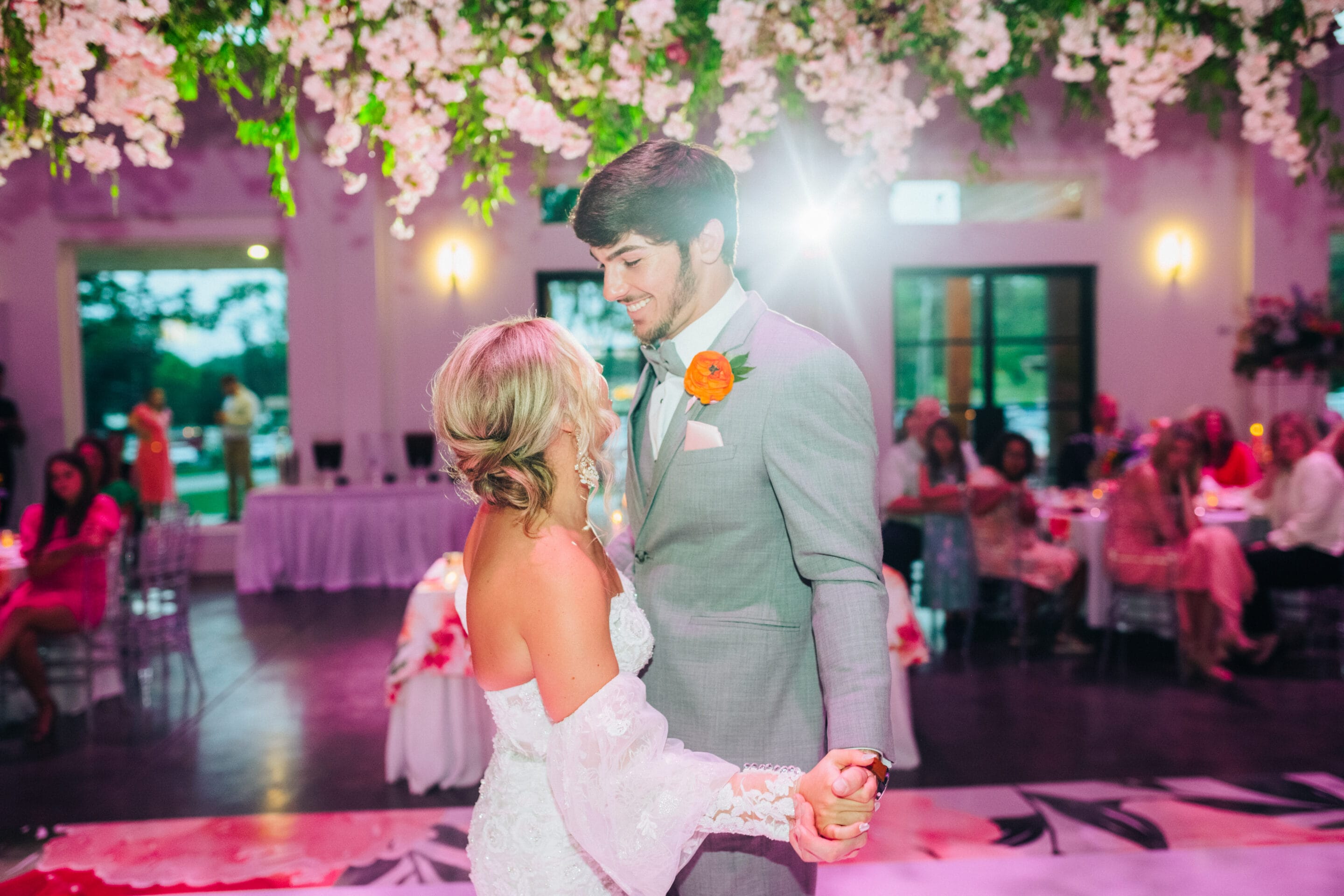 A bride and groom share a joyful first dance under hanging white flowers at their wedding reception, smiling at each other as guests watch from decorated tables in the background.