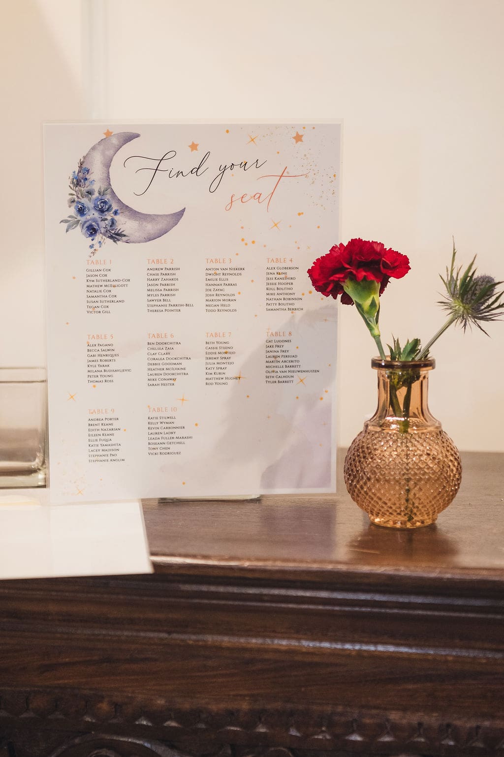 A “Find your seat” Eclipse Weekend Wedding seating chart with a crescent moon and blue flowers is displayed on a wooden table next to a textured bronze vase holding red carnations and greenery.