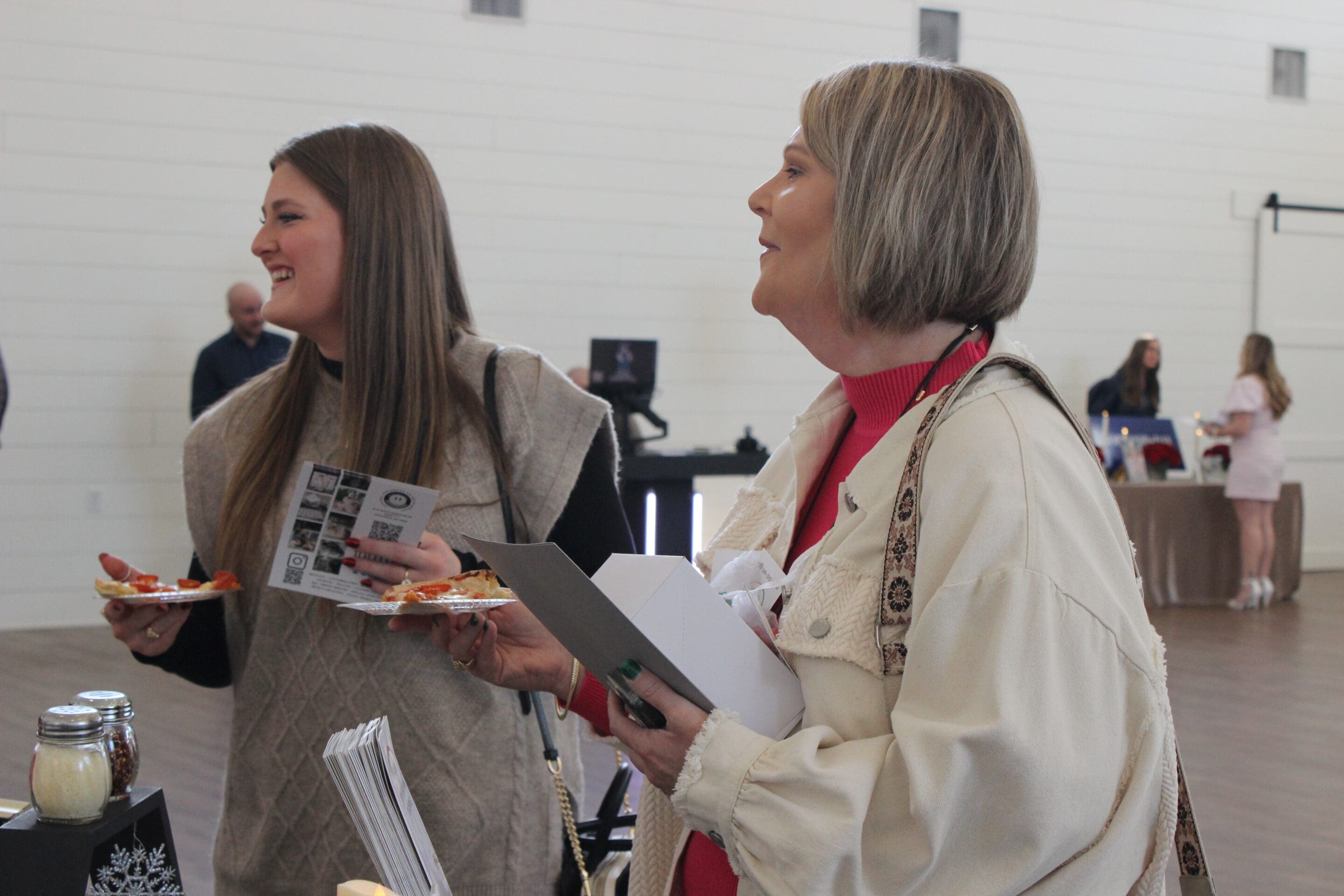 Two women stand indoors at the Married & Bright Wedding Event, holding food and brochures. They are smiling and appear to be engaged in conversation, with other people and display tables visible in the background.