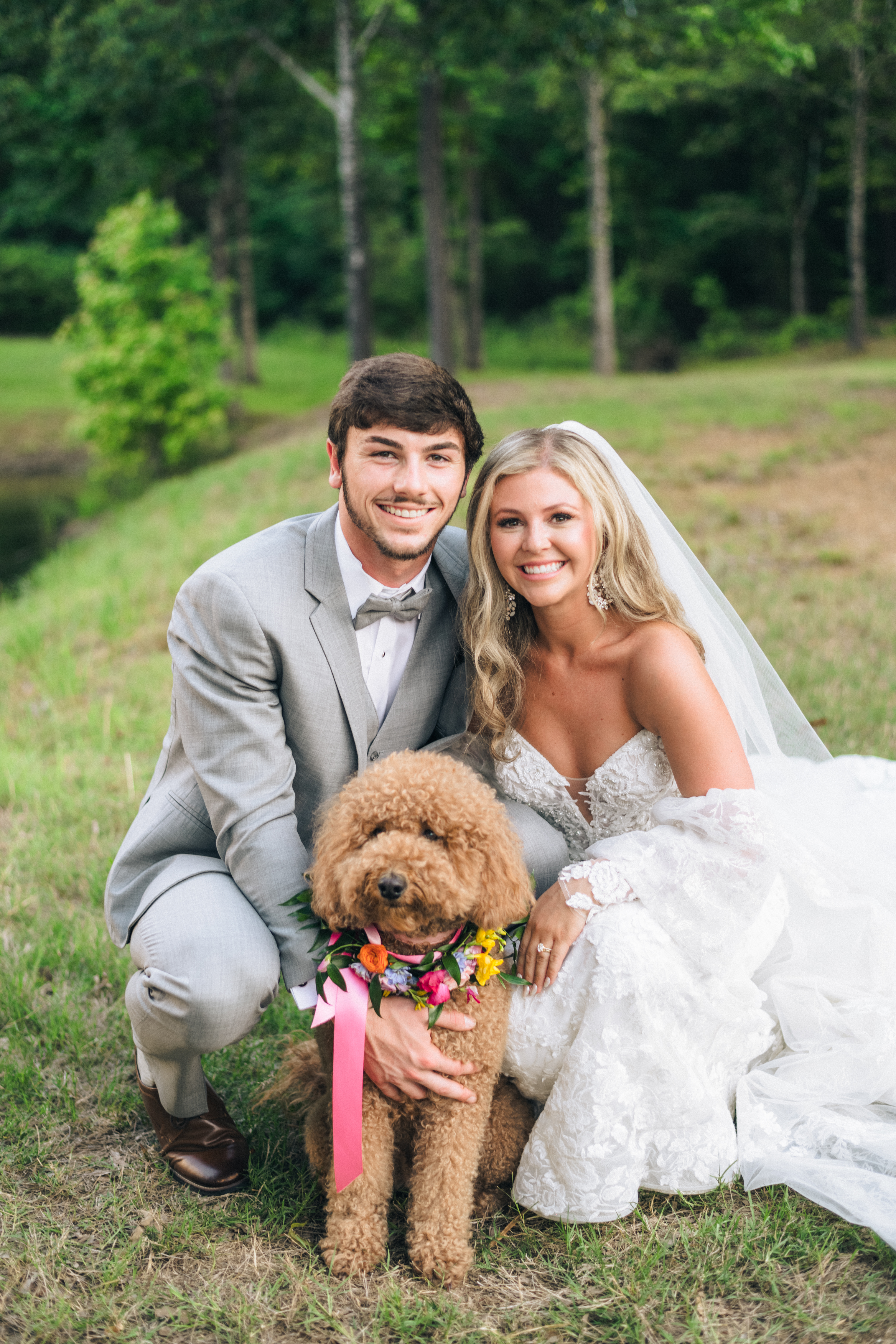 A bride and groom in wedding attire kneel outdoors on grass, smiling next to a fluffy brown dog wearing a colorful floral collar. Trees and greenery are in the background.