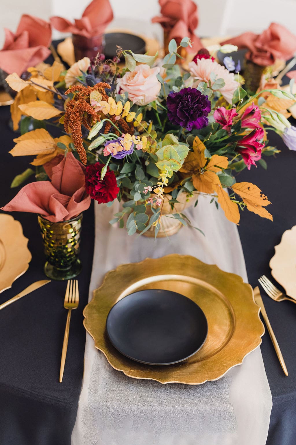 Elegant table setting for an Eclipse Weekend Wedding with a black tablecloth, gold plates, black bowls, gold cutlery, green glass, rust-colored napkins, and a vibrant floral centerpiece featuring autumn leaves and colorful flowers.