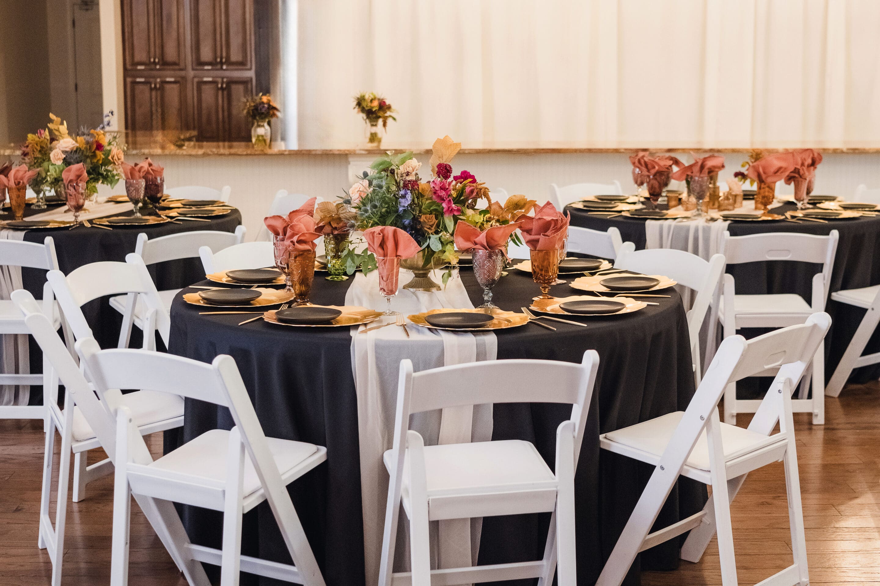 Round tables with black tablecloths, floral centerpieces, and peach napkins folded in glasses, surrounded by white chairs, create an elegant Eclipse Weekend Wedding setup in a well-lit banquet room with a wooden floor.