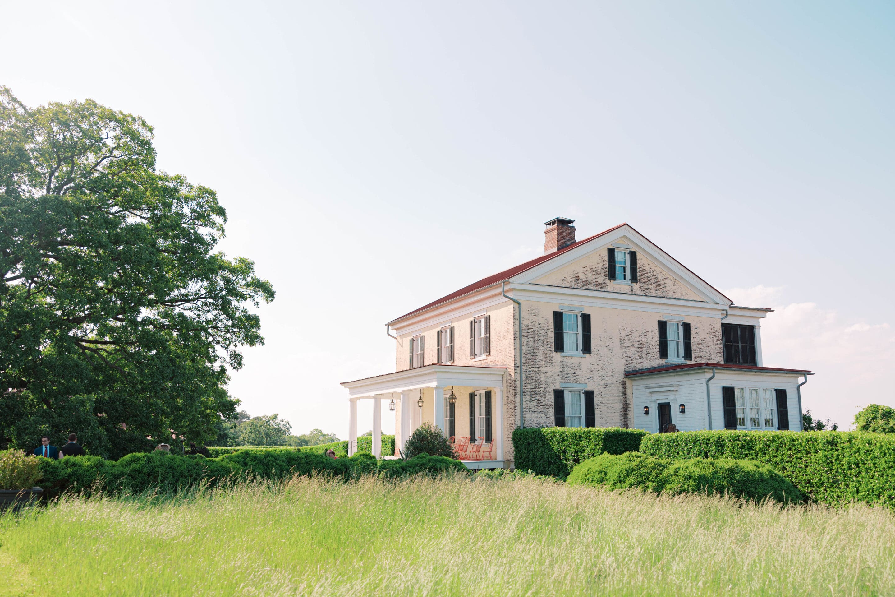 A large, two-story white house with black shutters sits amid tall green grass and manicured bushes on a sunny day, with a large tree to the left and clear blue sky in the background.