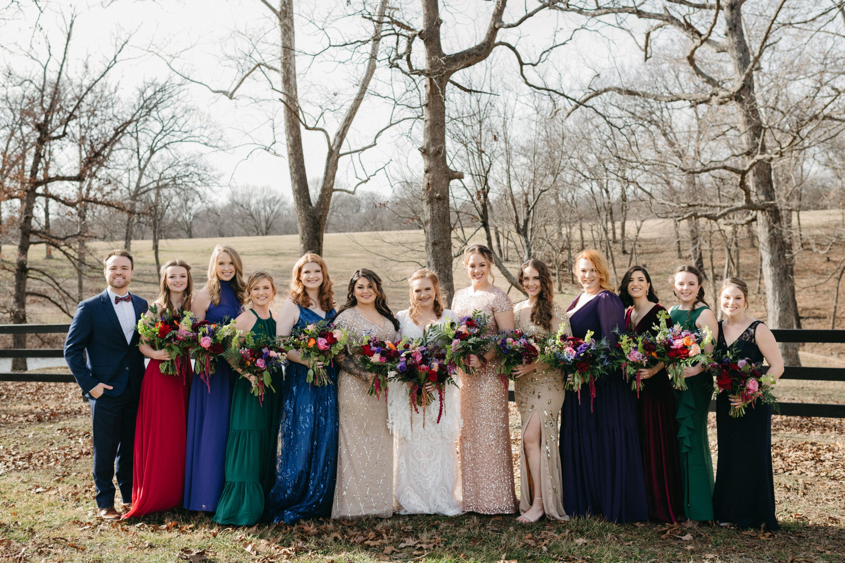 A wedding party stands outdoors in front of leafless trees and a wooden fence. The group includes one man in a suit and bow tie and thirteen women in colorful dresses, each holding a bouquet of flowers.