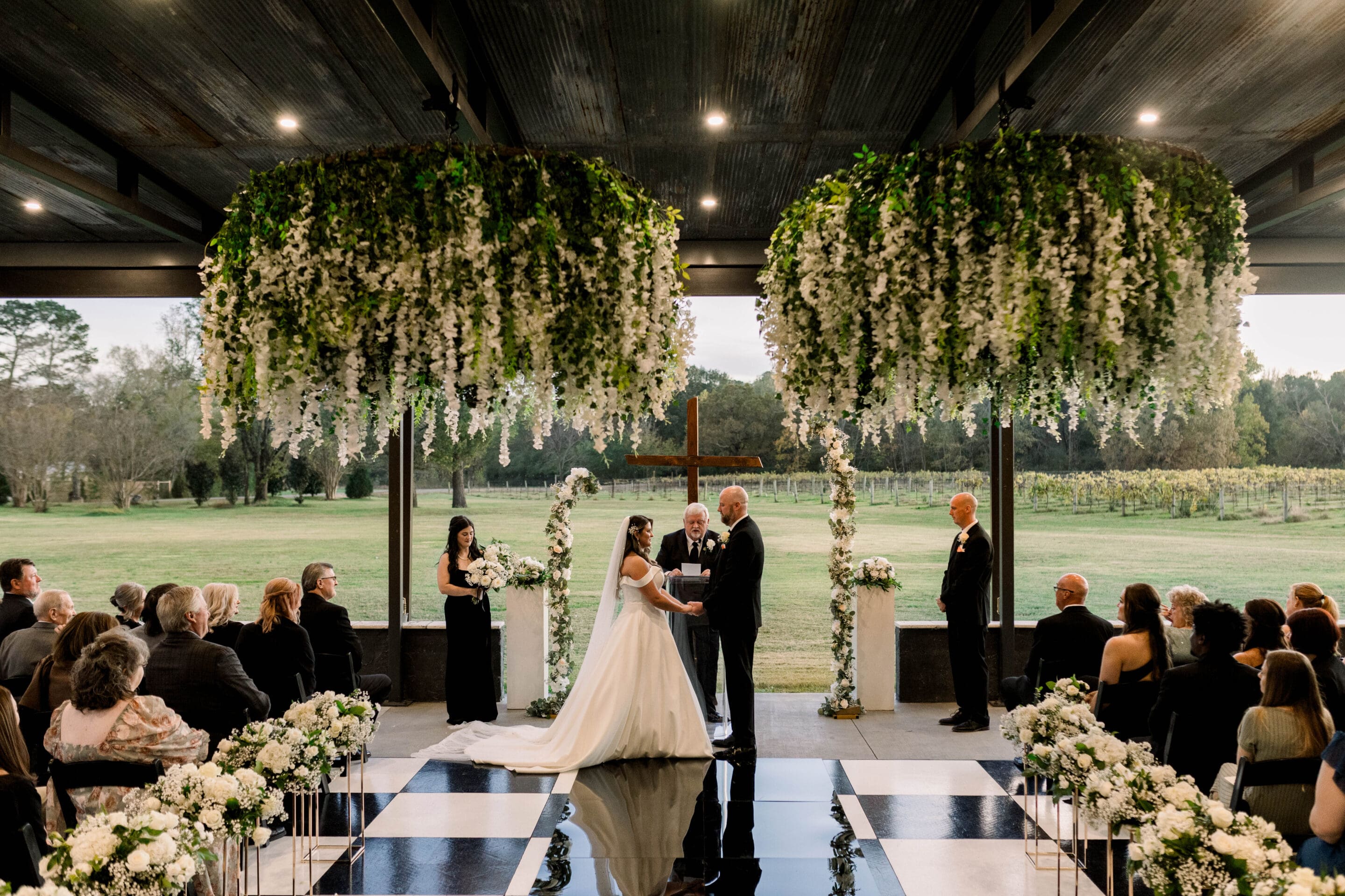 A bride and groom stand at the altar under hanging greenery, holding hands and facing each other during an indoor wedding ceremony with guests seated on both sides surrounded by white floral arrangements.