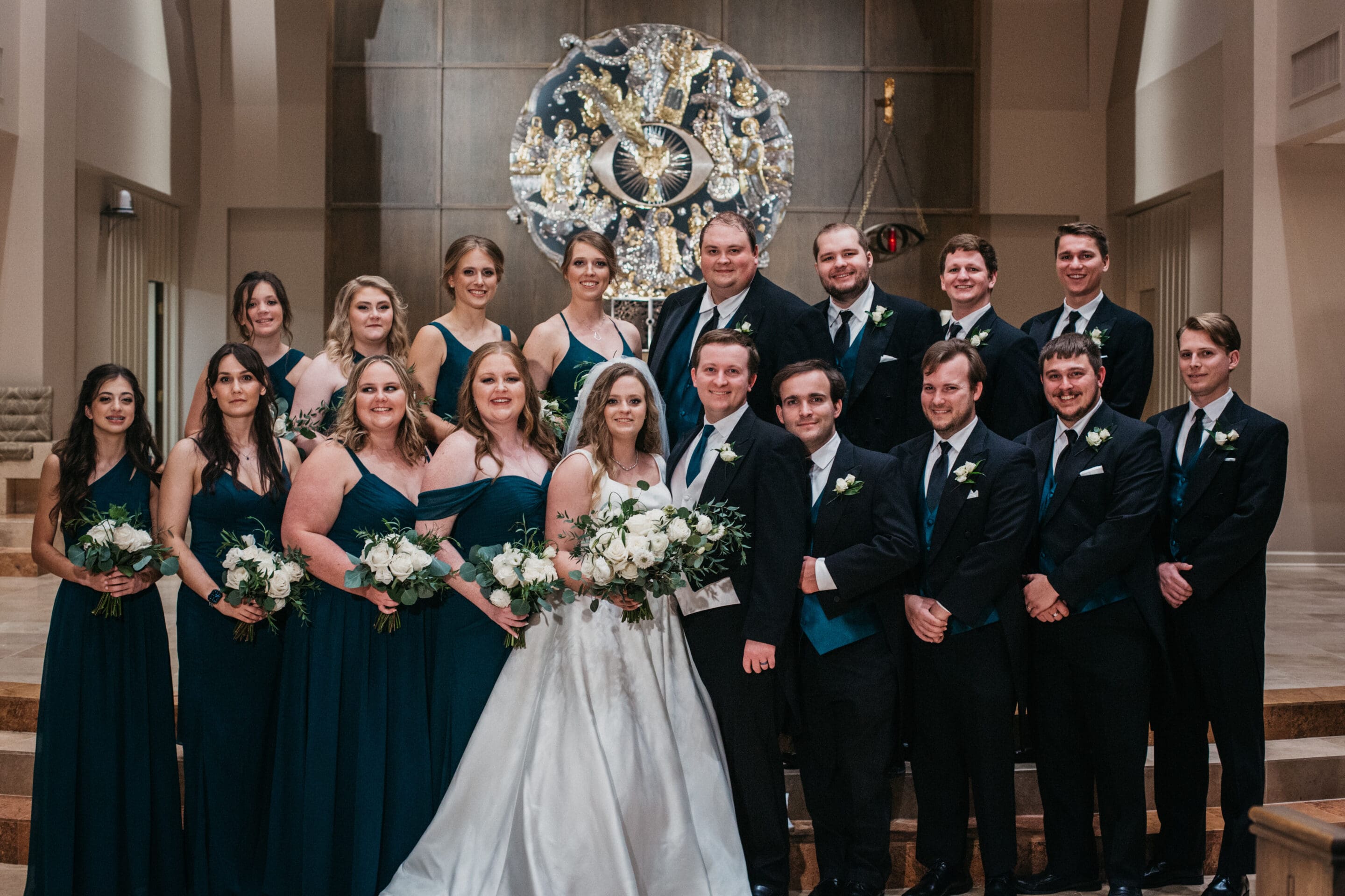 A wedding party poses indoors; the bride in a white gown stands center with the groom, surrounded by bridesmaids in dark green dresses and groomsmen in black suits, all holding bouquets or boutonnières, with ornate decor in the background.