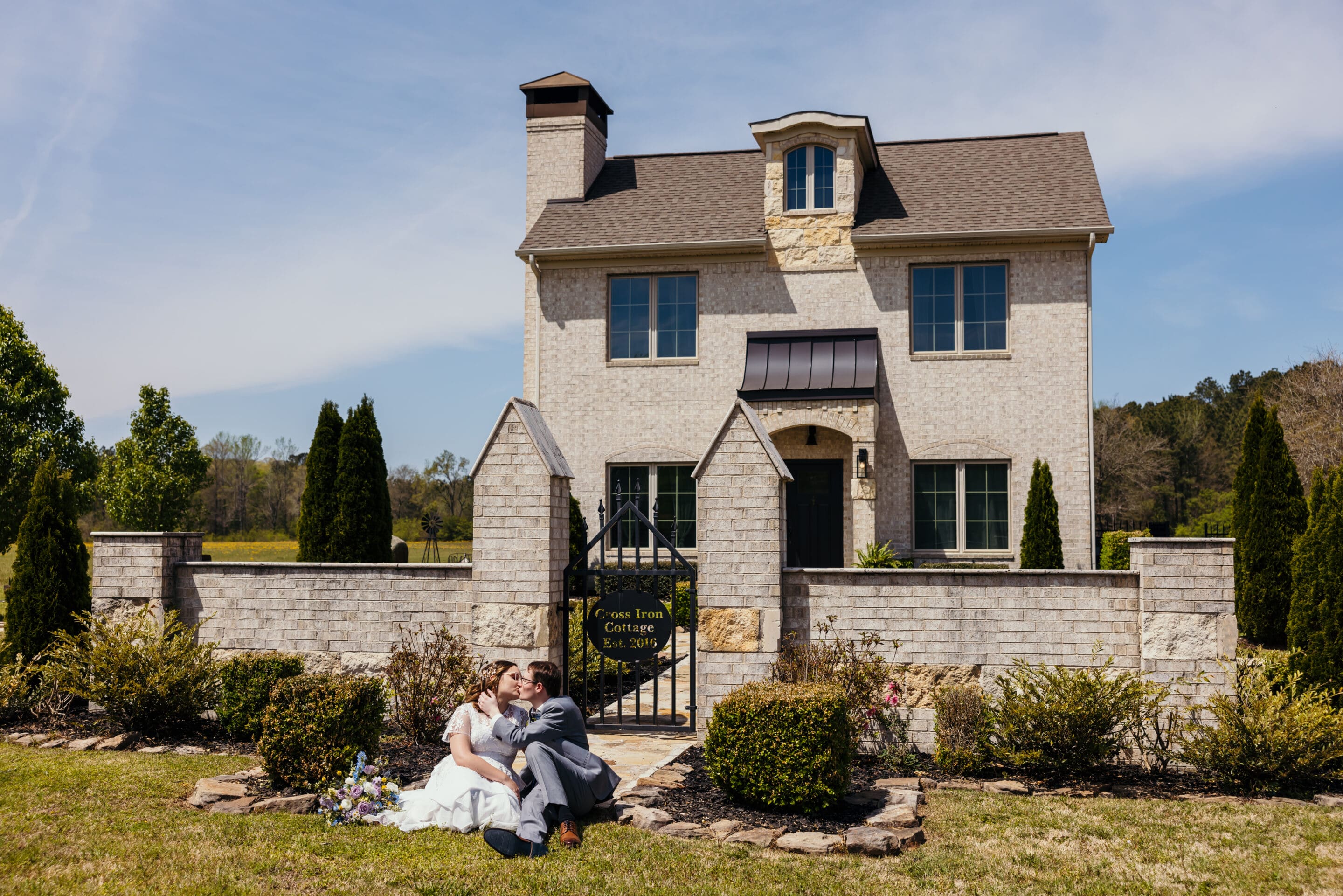 A bride and groom sit on the grass, embracing in front of a stone house with a gated entrance and a sign that reads Cove Home Cottage, Est. 2016. The day is sunny and the yard is landscaped with bushes and trees.
