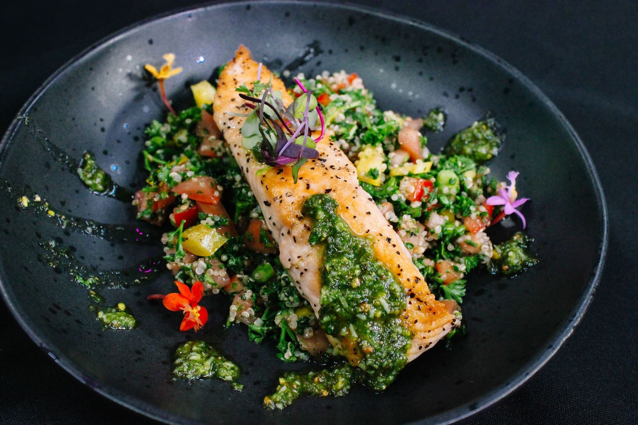 A grilled salmon fillet topped with green pesto, served on a bed of tabbouleh salad with diced tomatoes, cucumbers, and herbs, garnished with edible flowers, on a black plate.