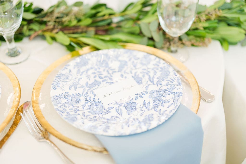 A blue and white floral-patterned plate with a name card rests on a gold charger atop a white tablecloth, accented by a light blue napkin and surrounded by greenery, a fork, and glassware.
