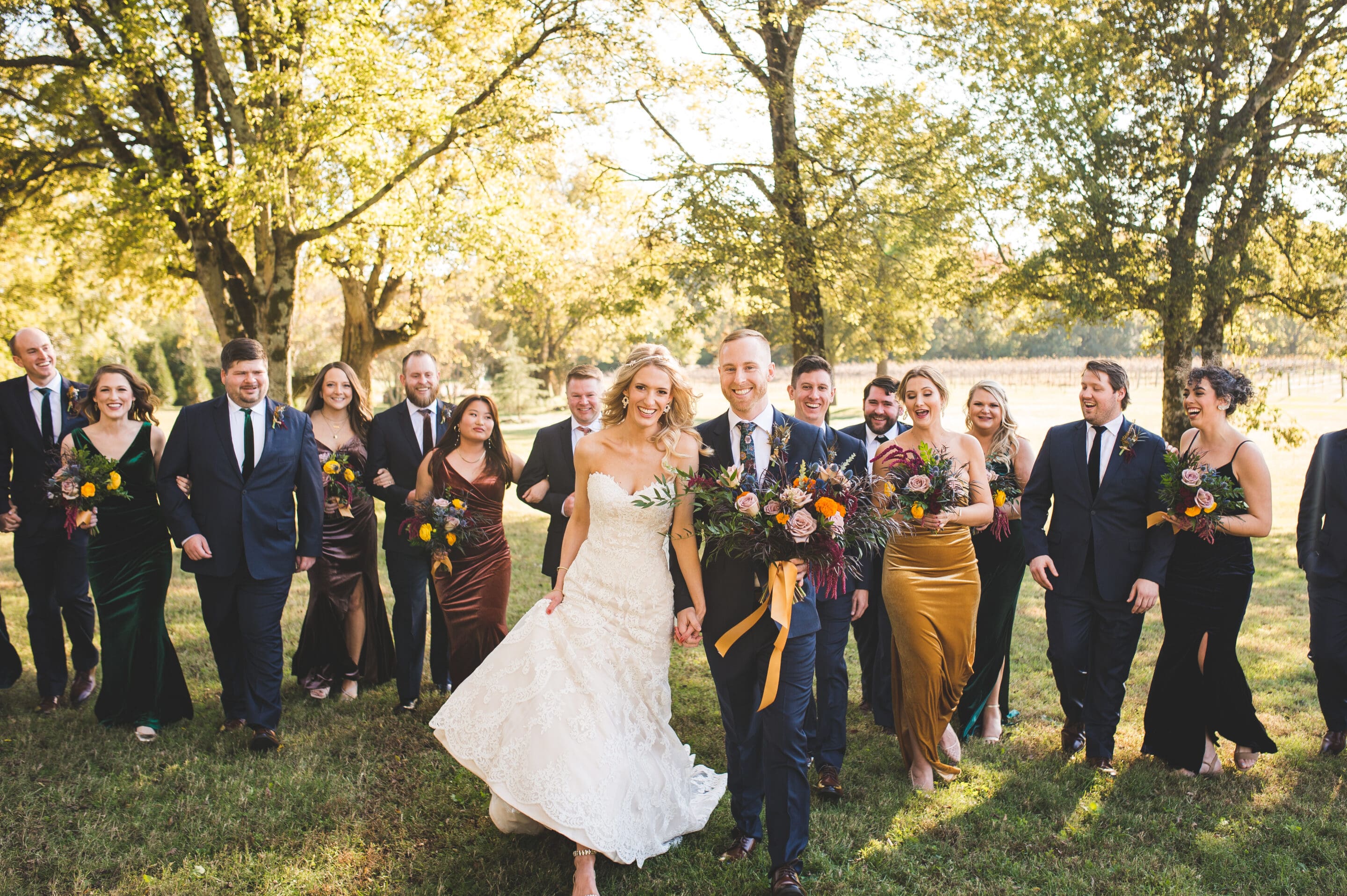 A bride and groom smile as they walk outside with their wedding party. Bridesmaids in colorful dresses and groomsmen in black suits surround them on a sunny, tree-filled lawn.