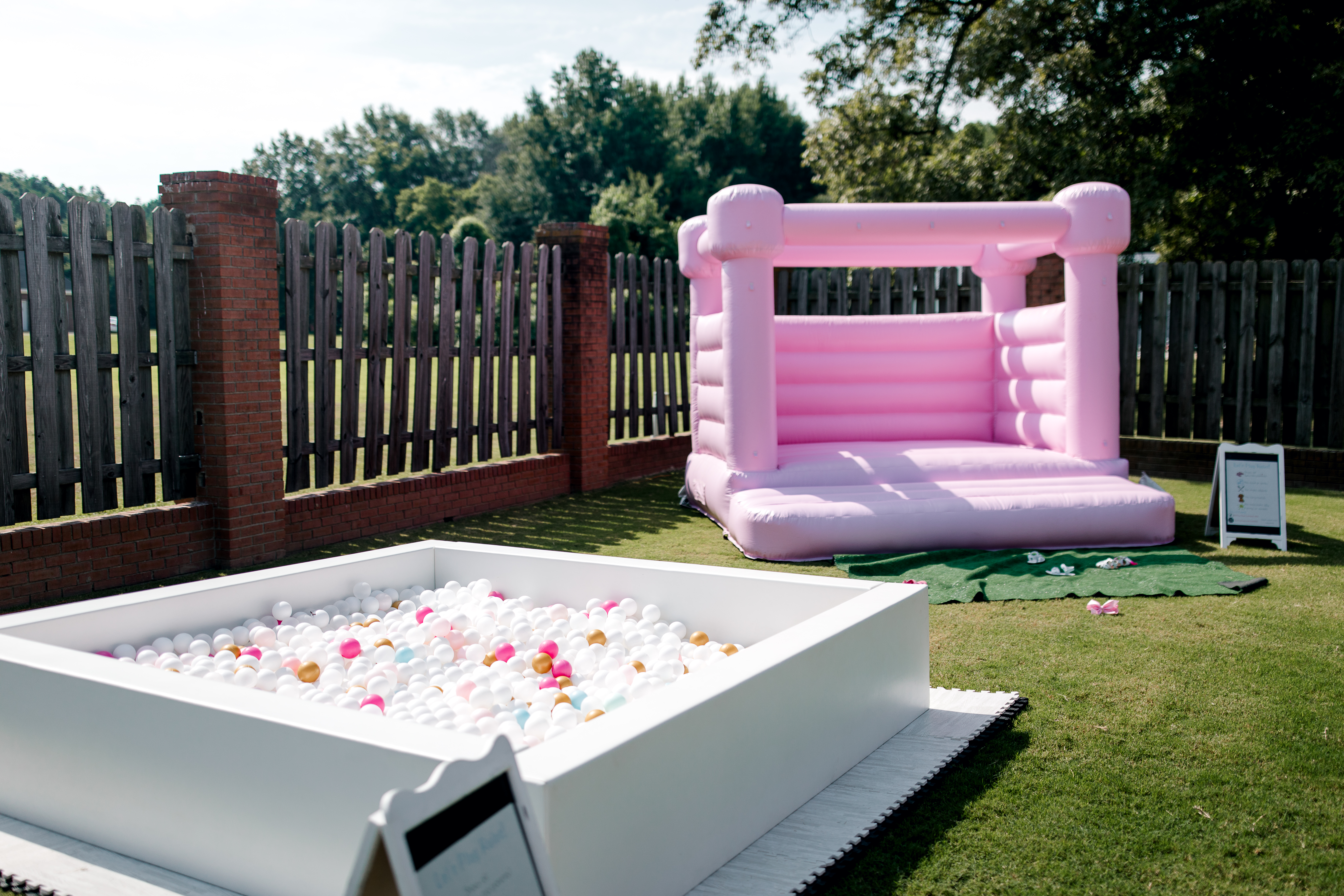 A pink inflatable bounce house and a white ball pit filled with colorful balls create the perfect setting for Poolside Princess Party Food, all arranged on grass near a wooden fence in an outdoor area on a sunny day.