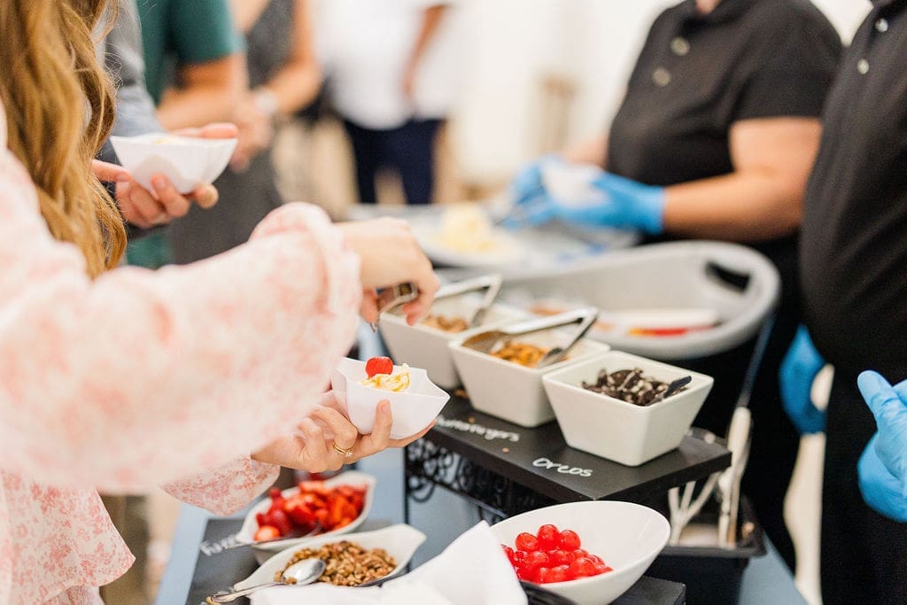 A person tops a bowl of dessert with cherry and spoon, selecting from a buffet of various toppings, while staff wearing black shirts and blue gloves assist in the background.