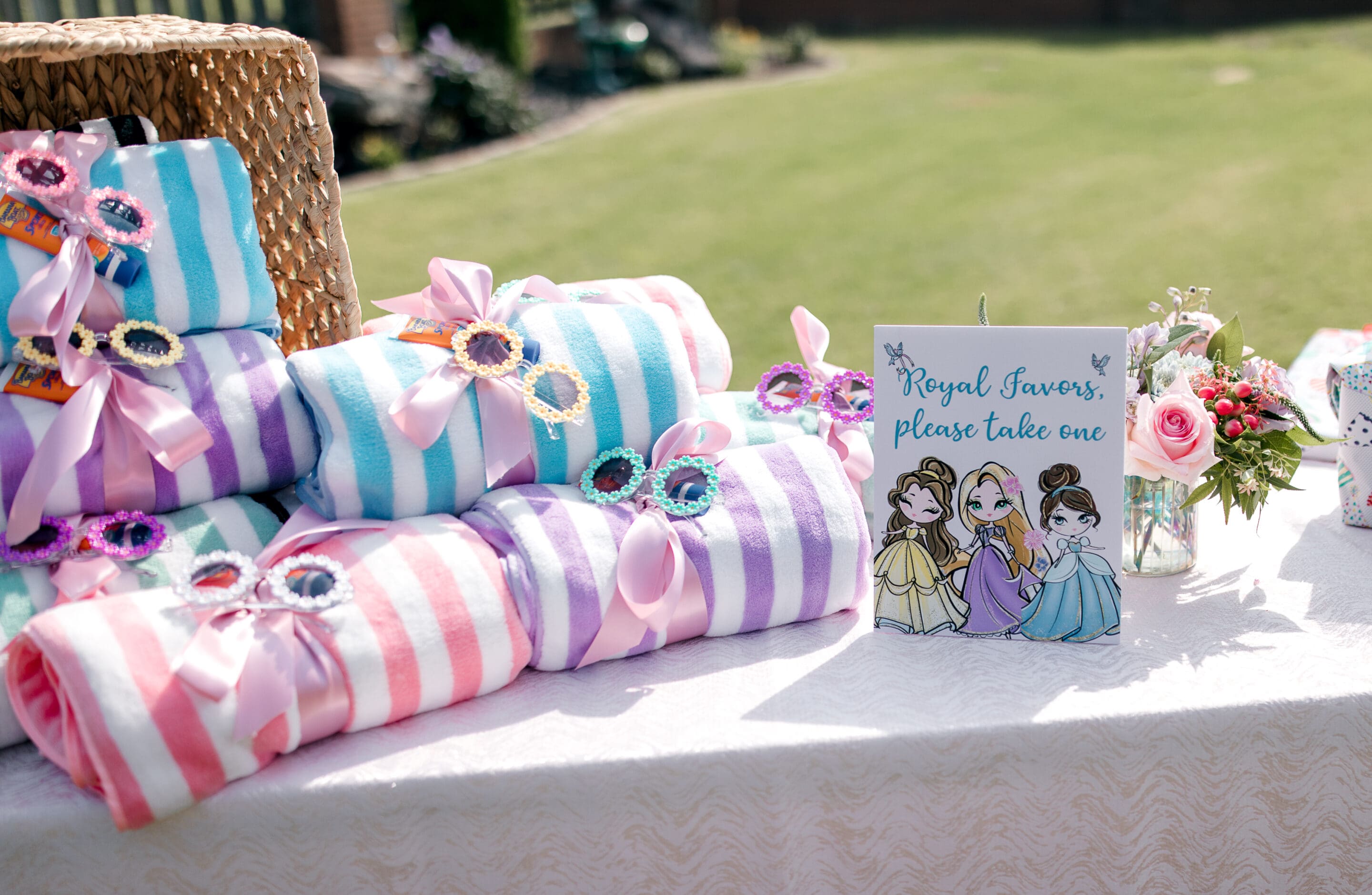 A table at the Poolside Princess Party displays pastel-striped towels tied with pink ribbon and toy princess tiaras. A sign with cartoon princesses reads, “Royal favors, please take one.” Flowers and a basket decorate the festive scene.