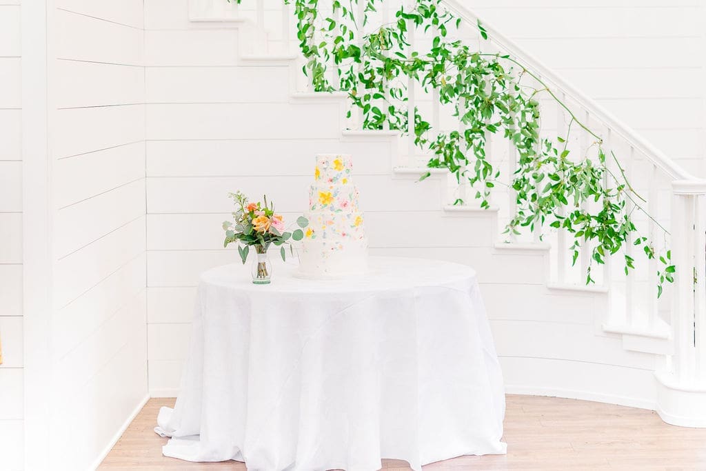 A three-tiered white cake with colorful floral decorations sits on a round table draped with a white cloth, next to a small bouquet. Green vines decorate a white staircase in the background.