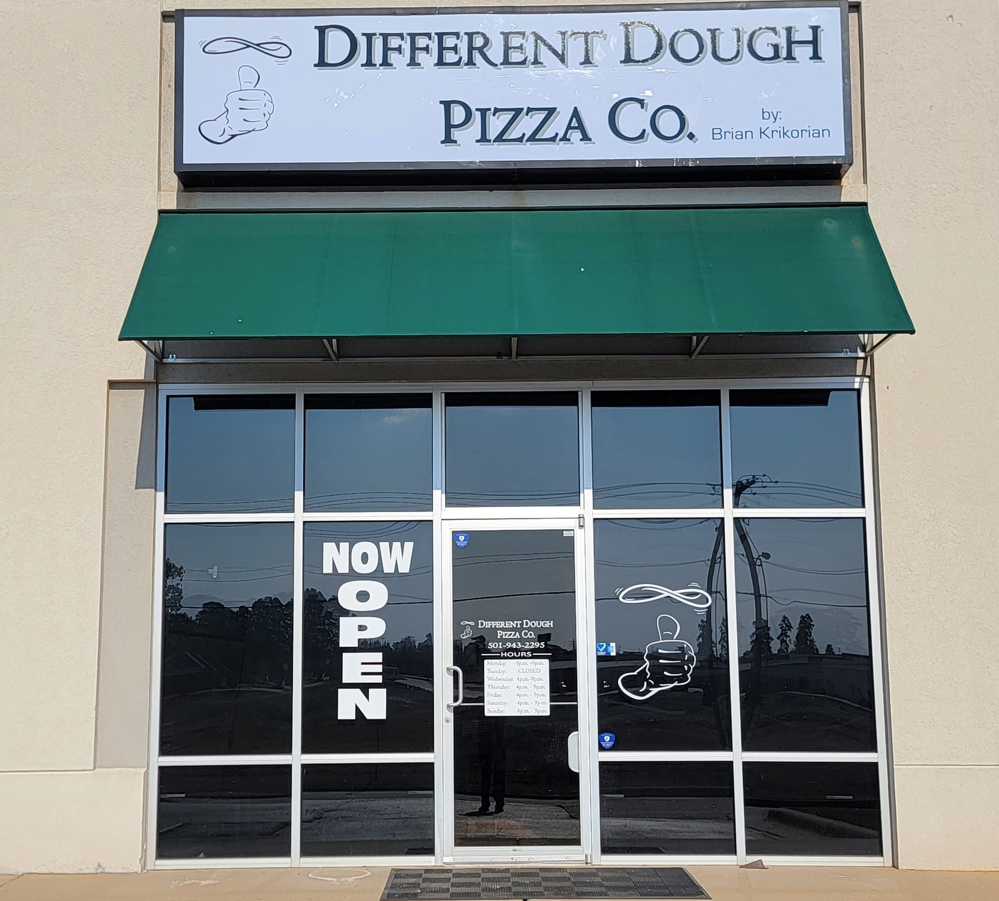 Storefront of “Different Dough Pizza Co.” with a green awning, large glass doors, and “NOW OPEN” signs displayed. The logo features a hand holding pizza dough. The area outside is sunny with a clear view of the street.