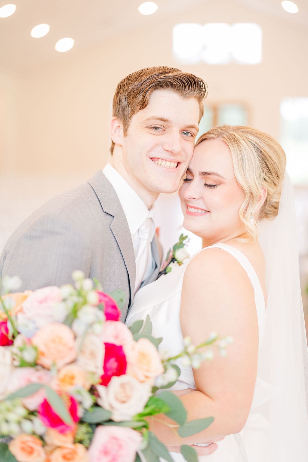 A smiling bride and groom embrace indoors, holding a colorful bouquet of roses and greenery. The bride wears a white dress and veil, eyes closed, while the groom wears a light gray suit and looks at the camera.