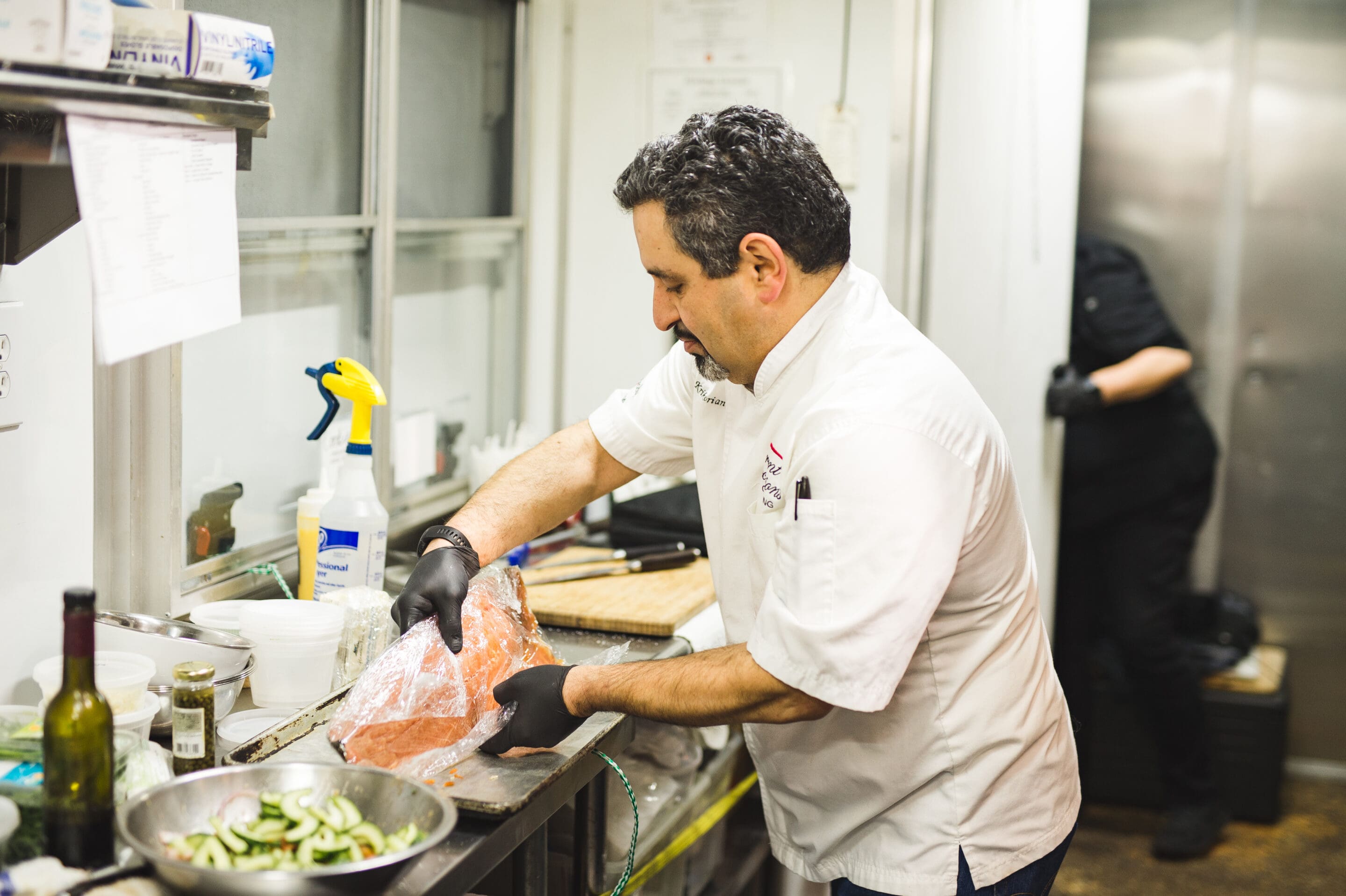 A chef wearing a white coat and black gloves prepares a large piece of salmon in a commercial kitchen, while another person works in the background. There are ingredients and utensils on the counter.