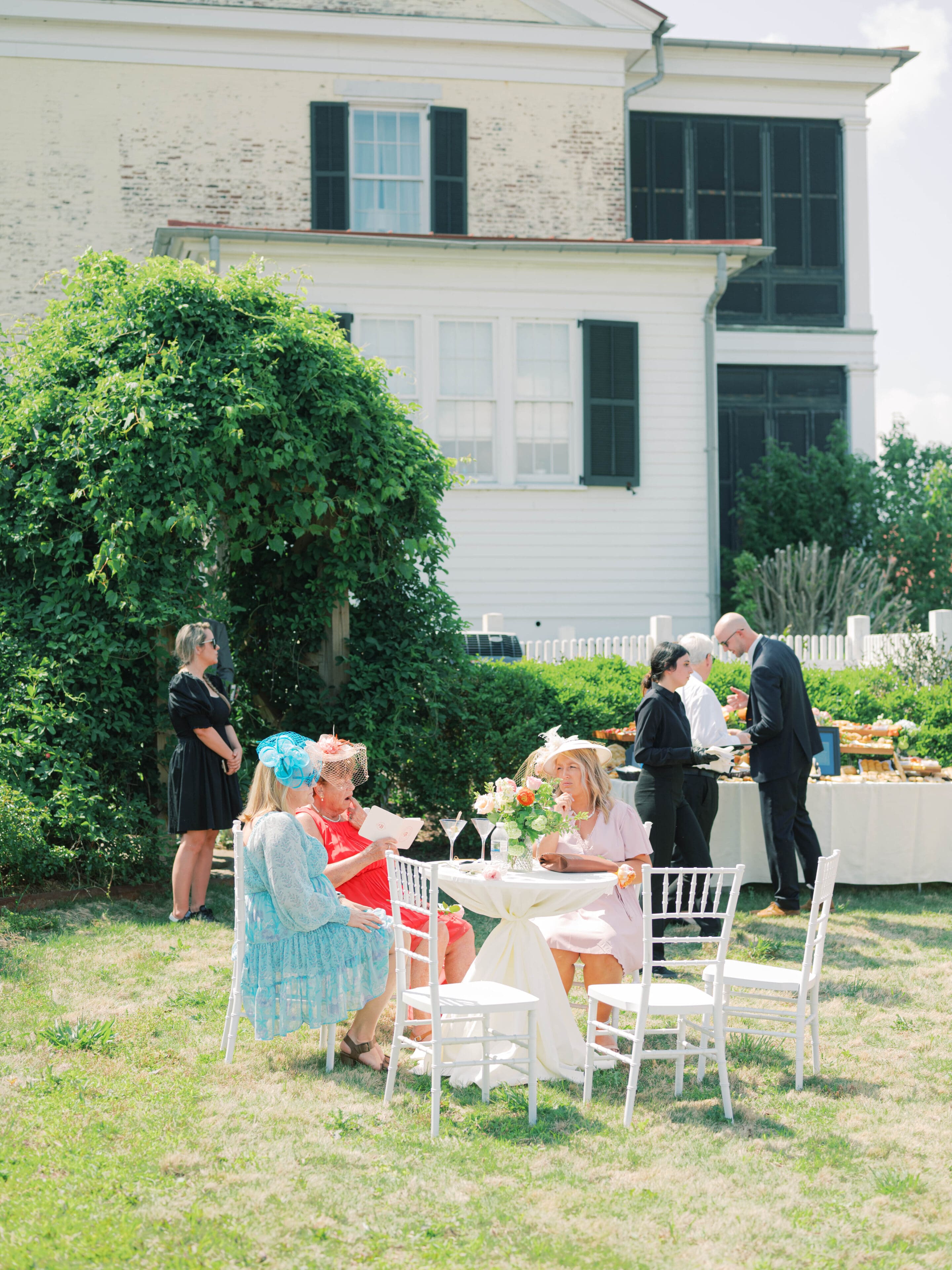 Three women in colorful dresses and hats sit at a round table outdoors, chatting and eating. Two servers in black stand nearby, with a buffet setup behind them. A large house and greenery are in the background.