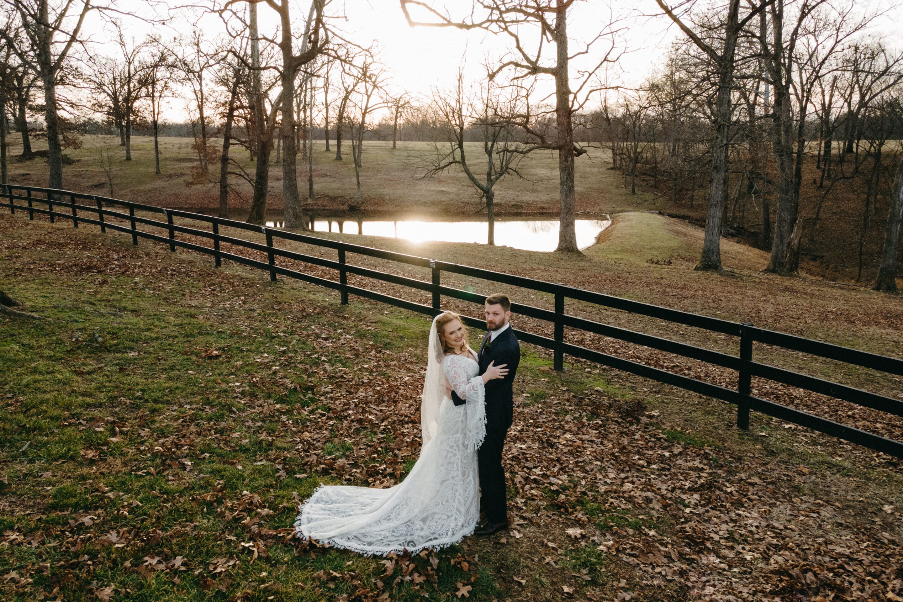 A bride and groom in wedding attire stand together on a grassy hill covered with autumn leaves, near a black wooden fence and a small pond, with bare trees and a sunset in the background.