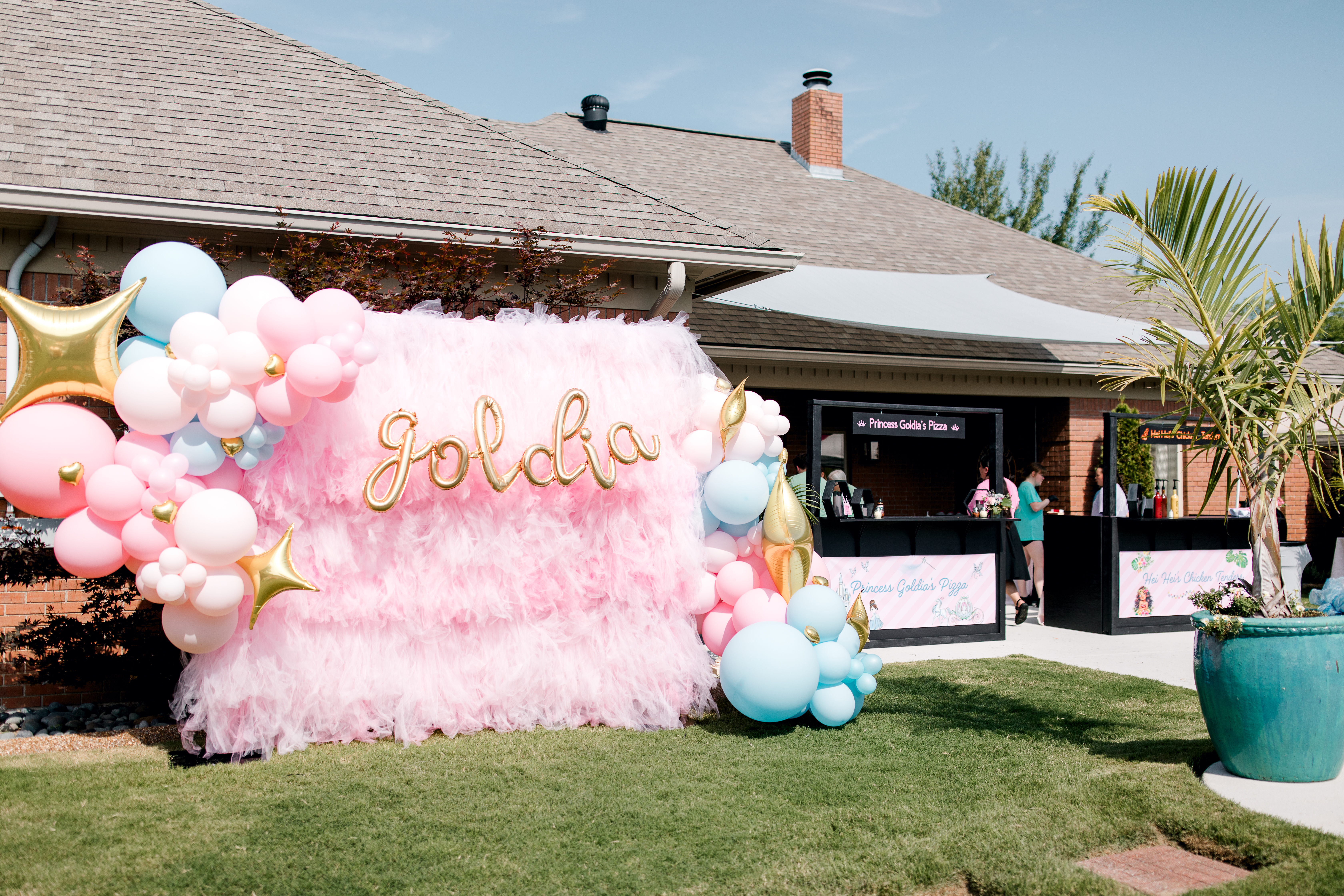 The image shows an outdoor celebration with a pink and white balloon arch featuring the gold cursive name "Goldie" against a shimmery pink backdrop, set up on a green lawn near a house. Additional balloons and party decorations are visible, suggesting a festive event.