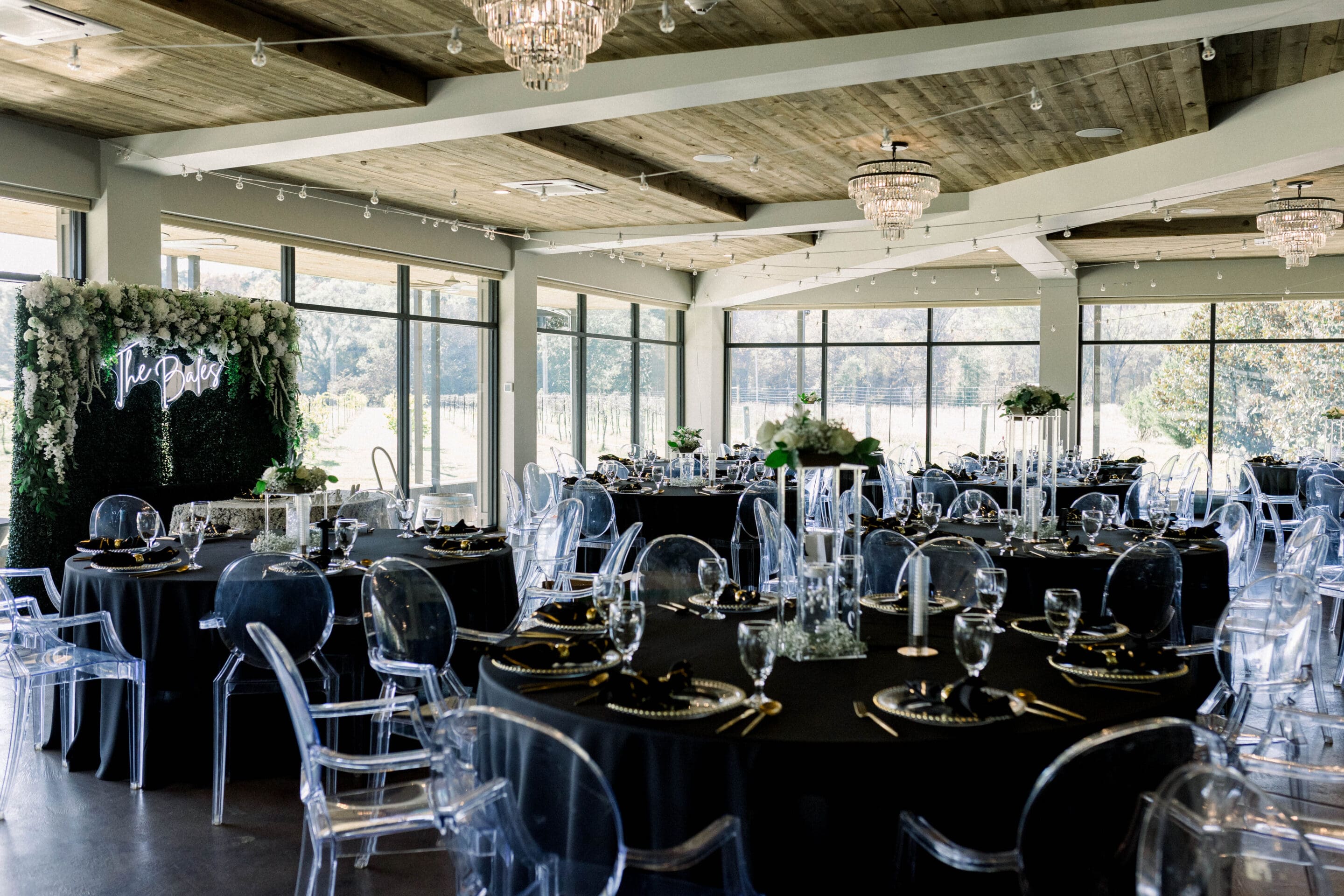 Elegant event venue with round tables covered in black tablecloths, clear acrylic chairs, floral arrangements, glassware, and a flower-covered backdrop with a neon sign. Large windows let in natural light.