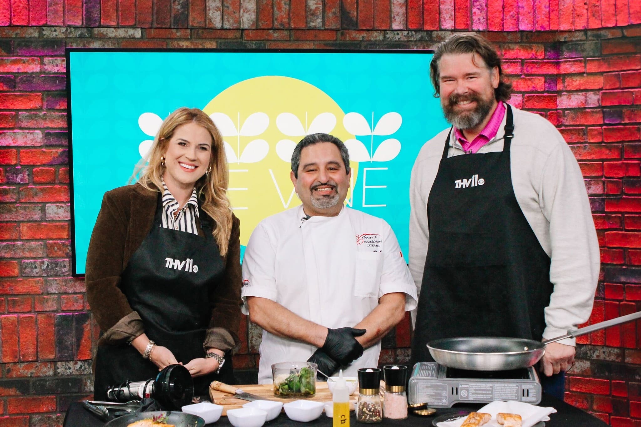 Three people stand smiling in a kitchen studio. The two on the left and right wear black aprons, while the person in the center wears a chefs uniform. Cooking ingredients and utensils are on the counter in front of them.