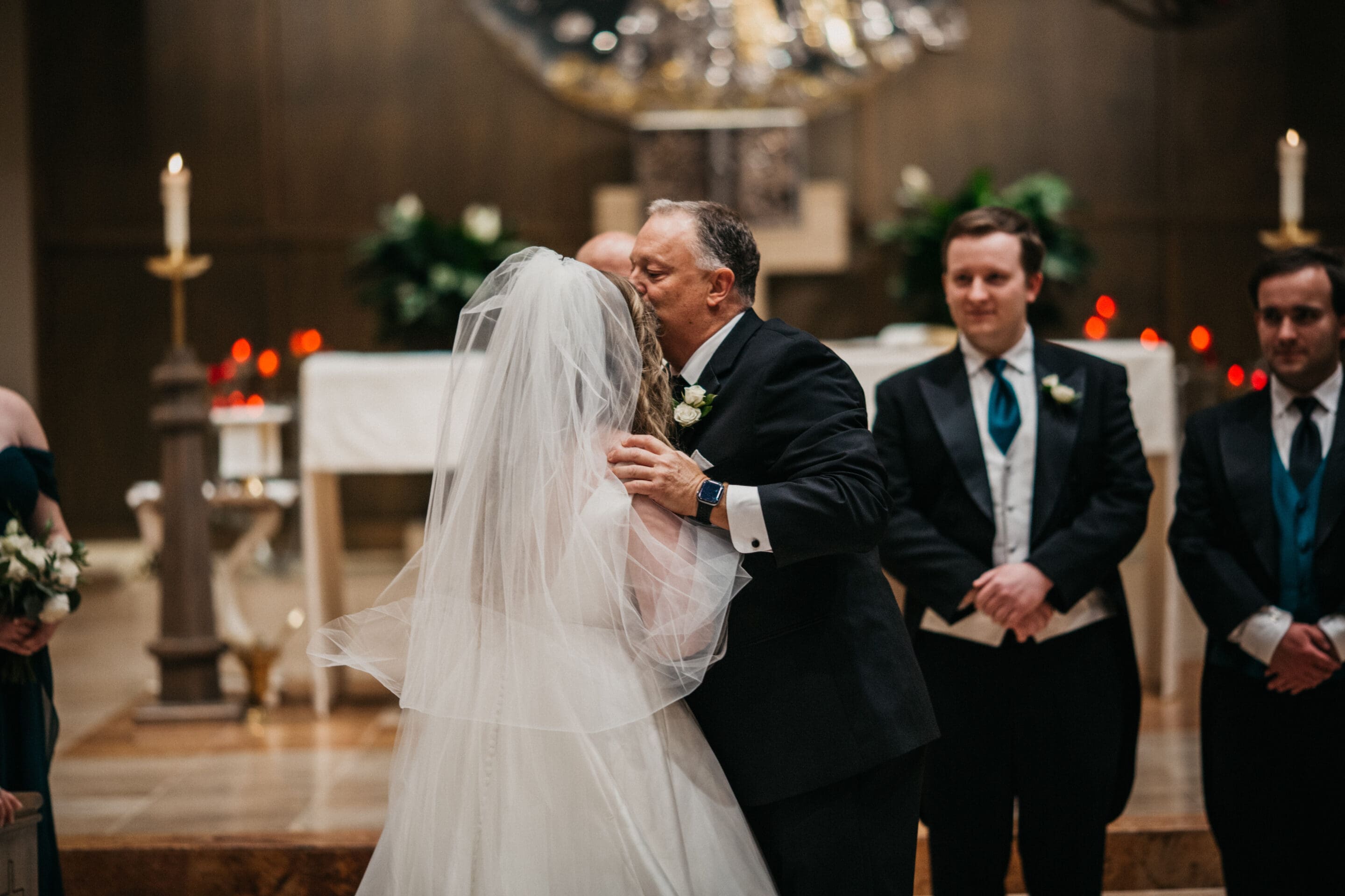 A man in a suit kisses a bride on the cheek at the altar, her face covered by a veil. Two groomsmen in tuxedos watch, and candles and floral arrangements decorate the background.